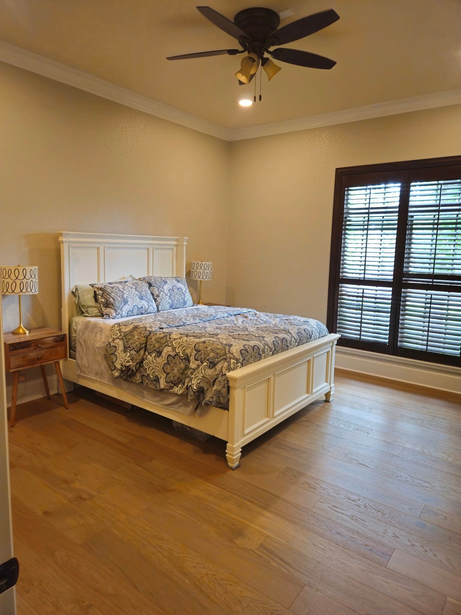 Bedroom with a white bed, floral bedding, wooden floor, and a dark-framed window with blinds.