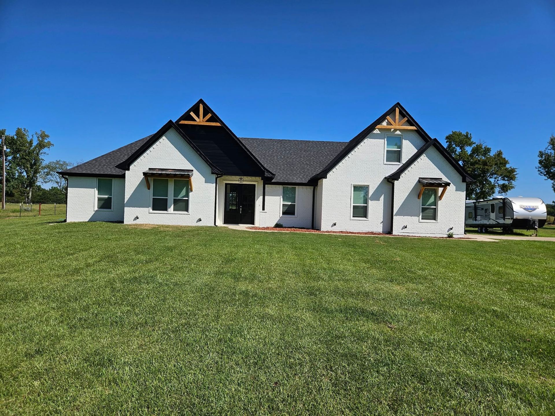 White brick house with black roof, wooden accents, and a trailer on the right.