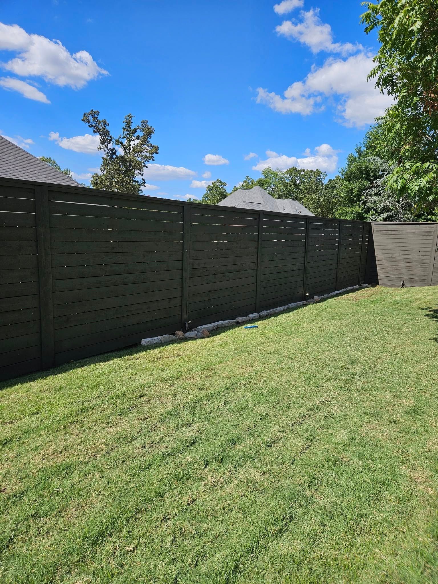 Black wooden fence in a grassy backyard under a blue sky with some trees.