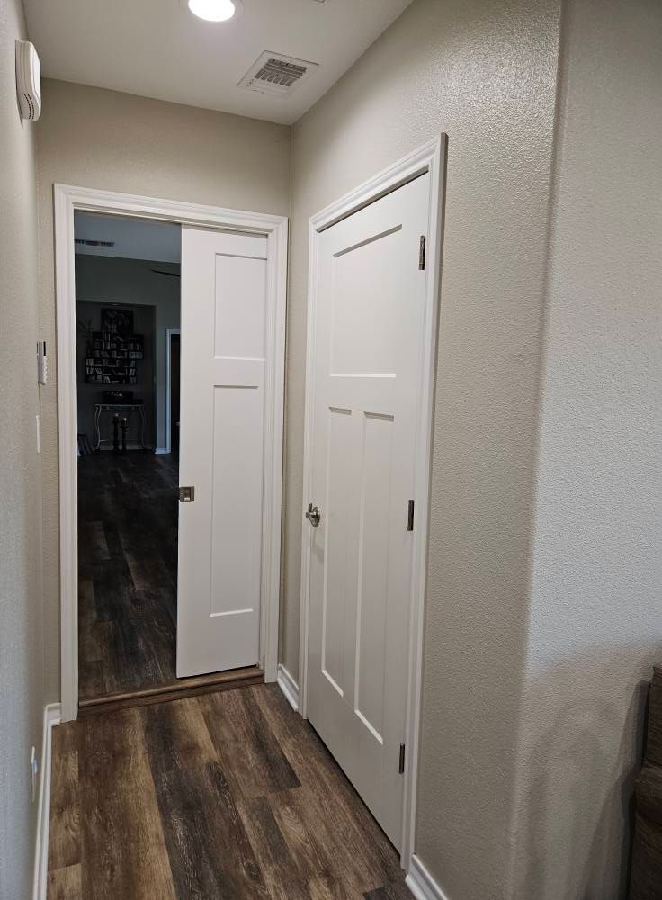 Hallway with two white doors, one open leading to a dark room, and wood-look flooring.