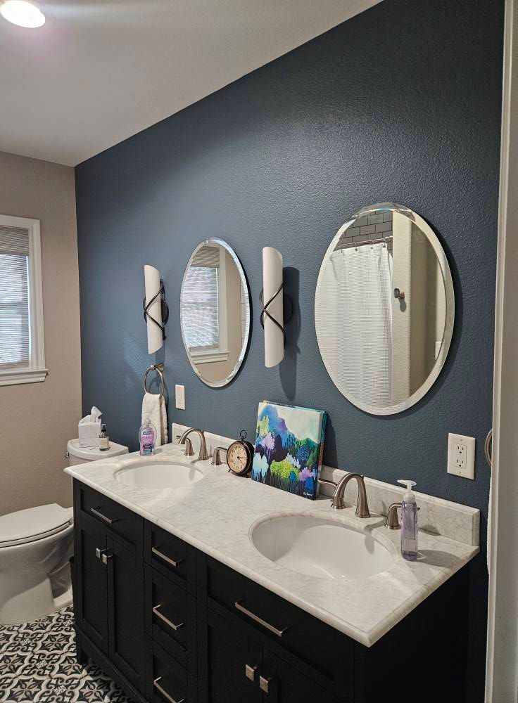 Bathroom with dark blue wall, double vanity, oval mirrors, and patterned floor.