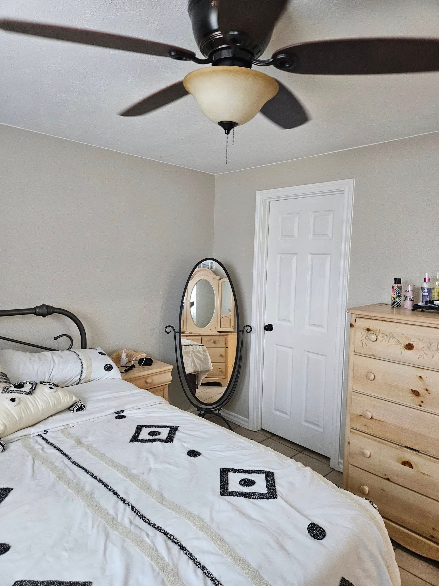 Bedroom with bed, mirror, chest of drawers, white door, and ceiling fan. The walls are beige.