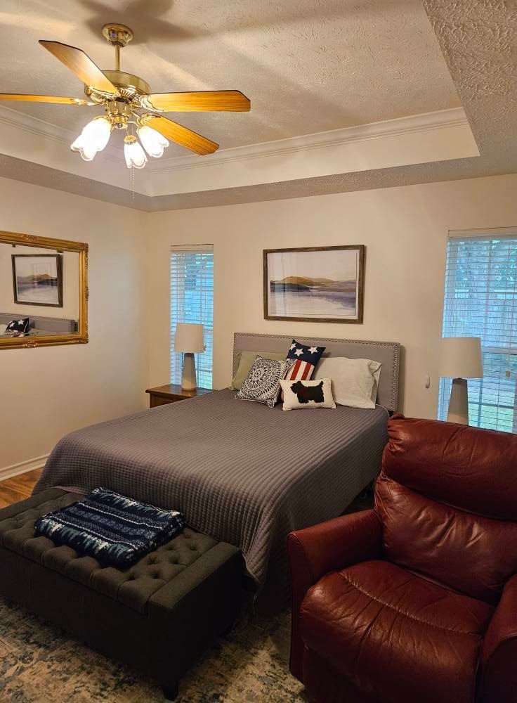 Bedroom with gray bed, red recliner, and decorative pillows. Ceiling fan and framed artwork.