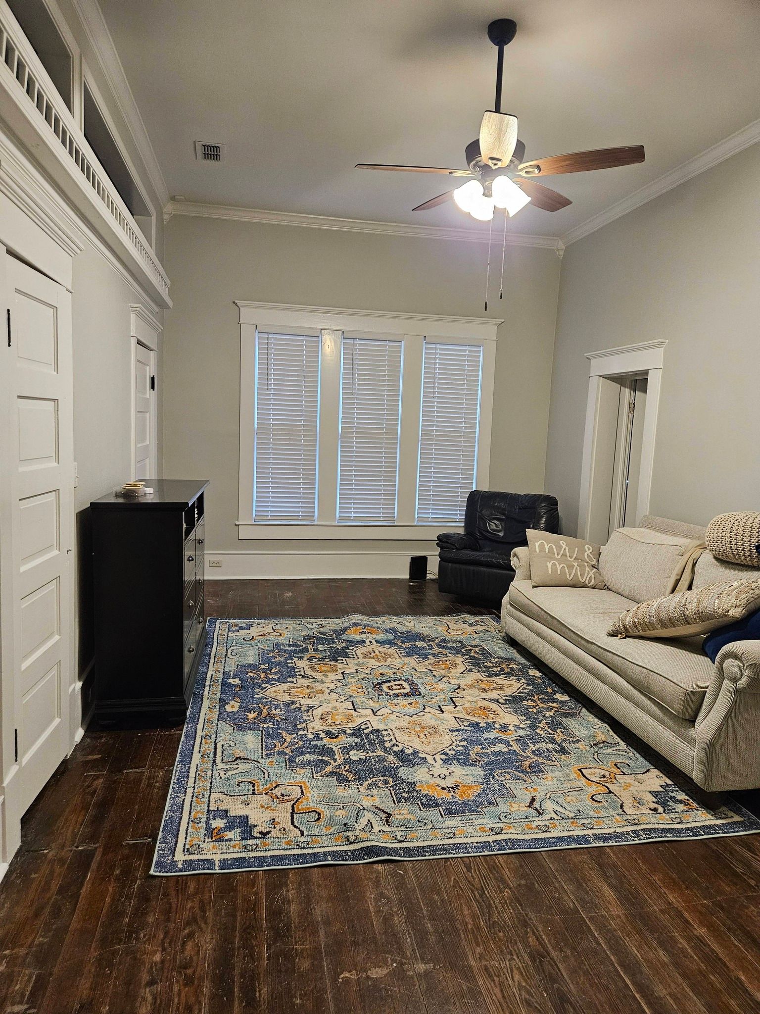 Living room with blue patterned rug, white couch, shutters, dark wood floors.