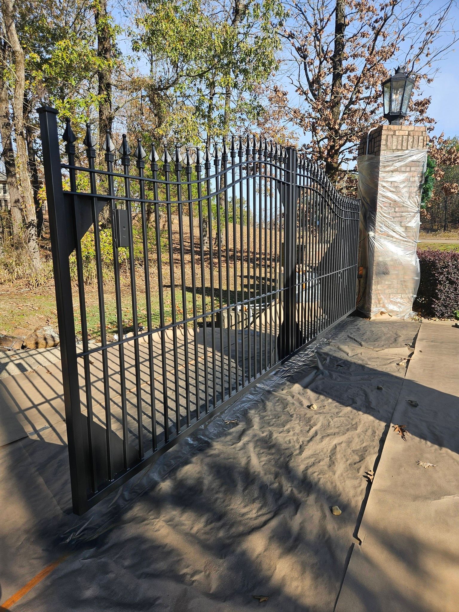 Black metal gate set into a cement path. Surrounding trees and a stone pillar.
