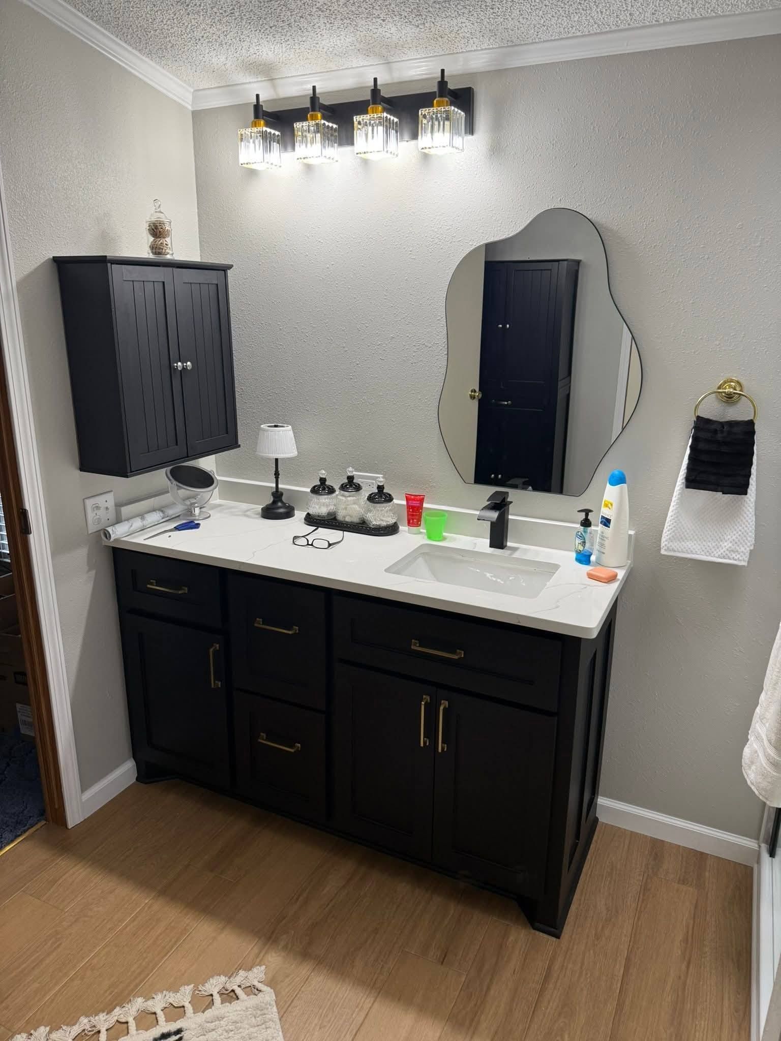 Black vanity with white countertop and wavy mirror in a bathroom with tan flooring. Black wall cabinet above.