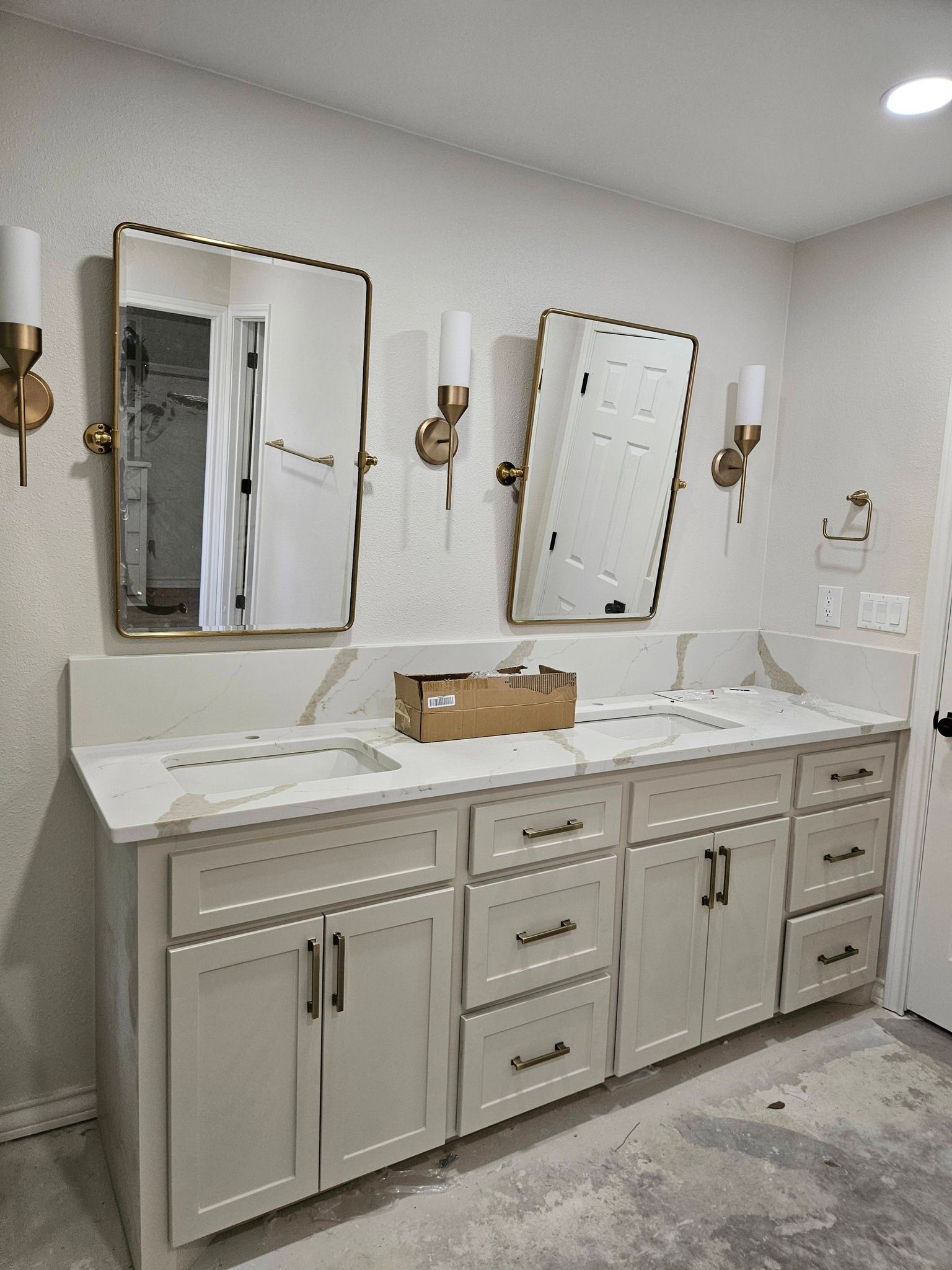 Bathroom vanity with two mirrors, lights, and cabinets. Light-colored countertop, white walls, and gold fixtures.