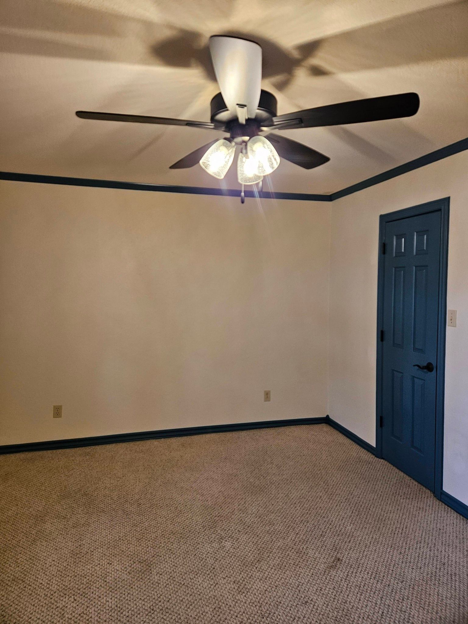 Empty room with tan carpet, white walls, and a teal door and trim. A ceiling fan hangs from the ceiling.