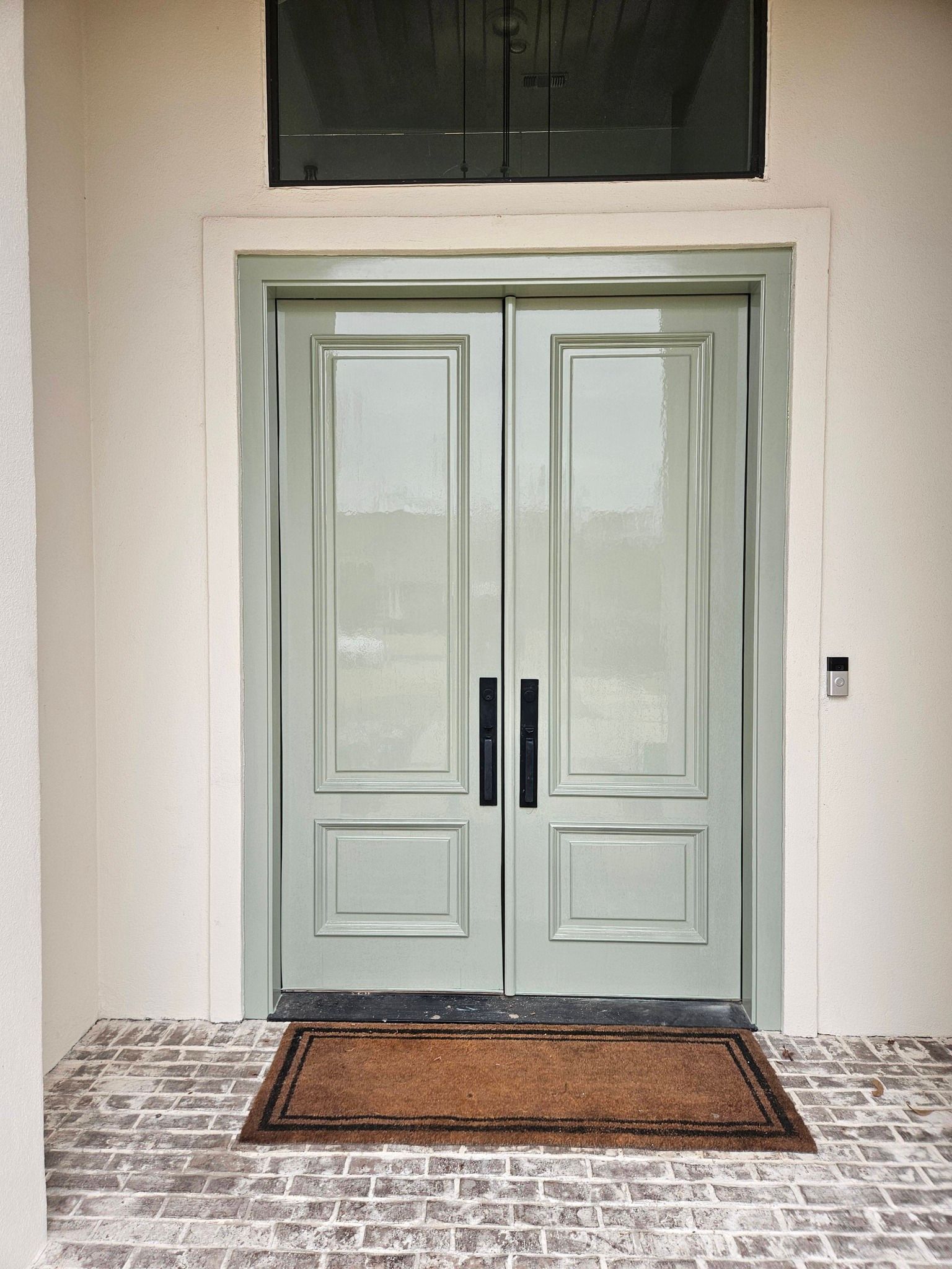 Double teal doors with rectangular glass panels, black handles, and brown welcome mat. Brick floor.