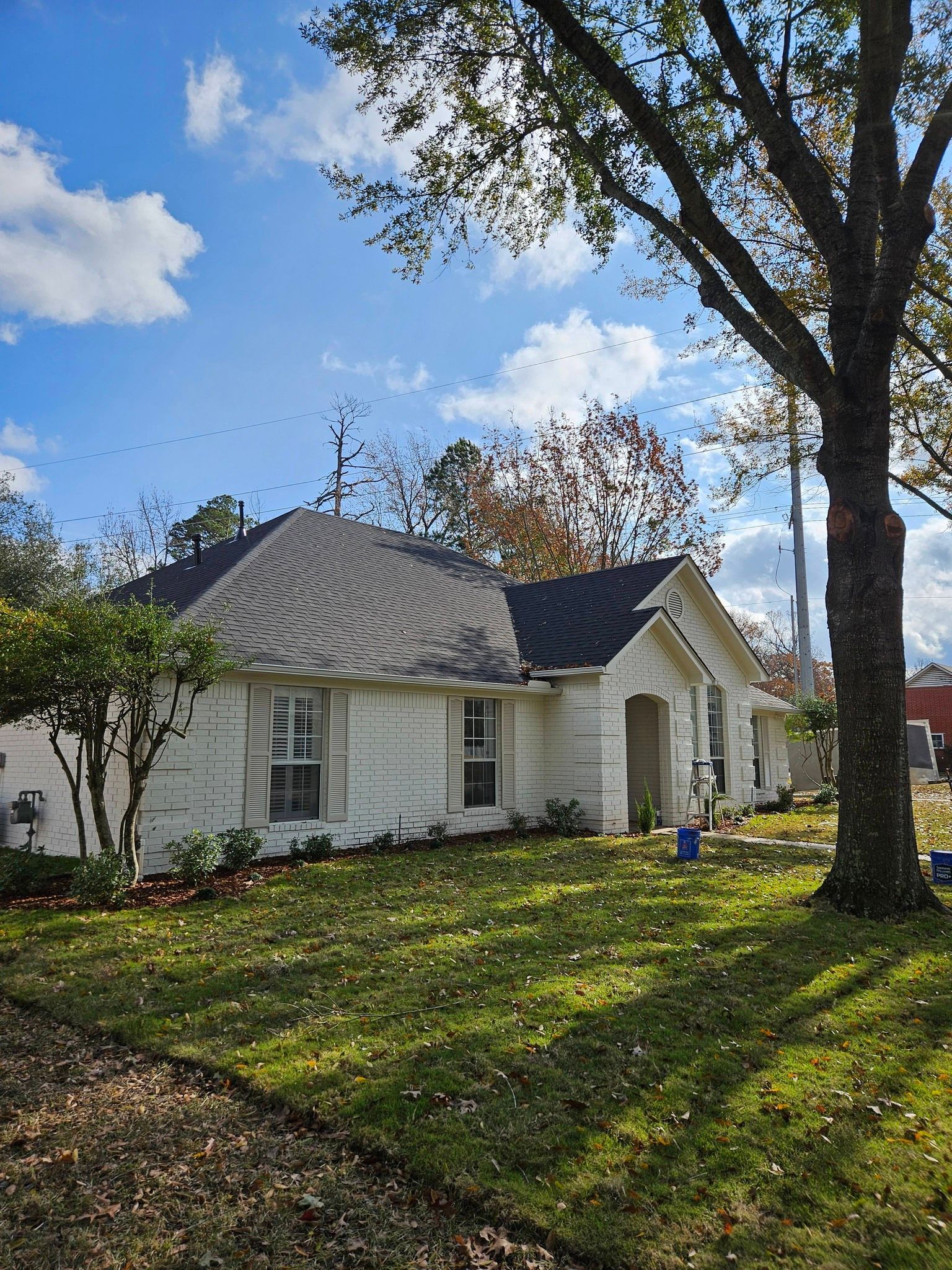 White house with dark roof and green lawn under a blue sky, shaded by trees.