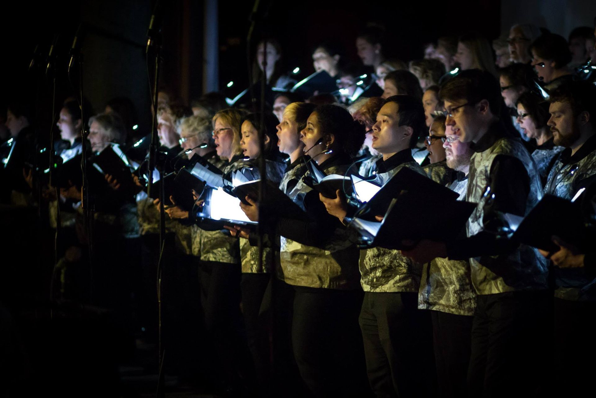 A large group of people are singing in a choir in a dark room.