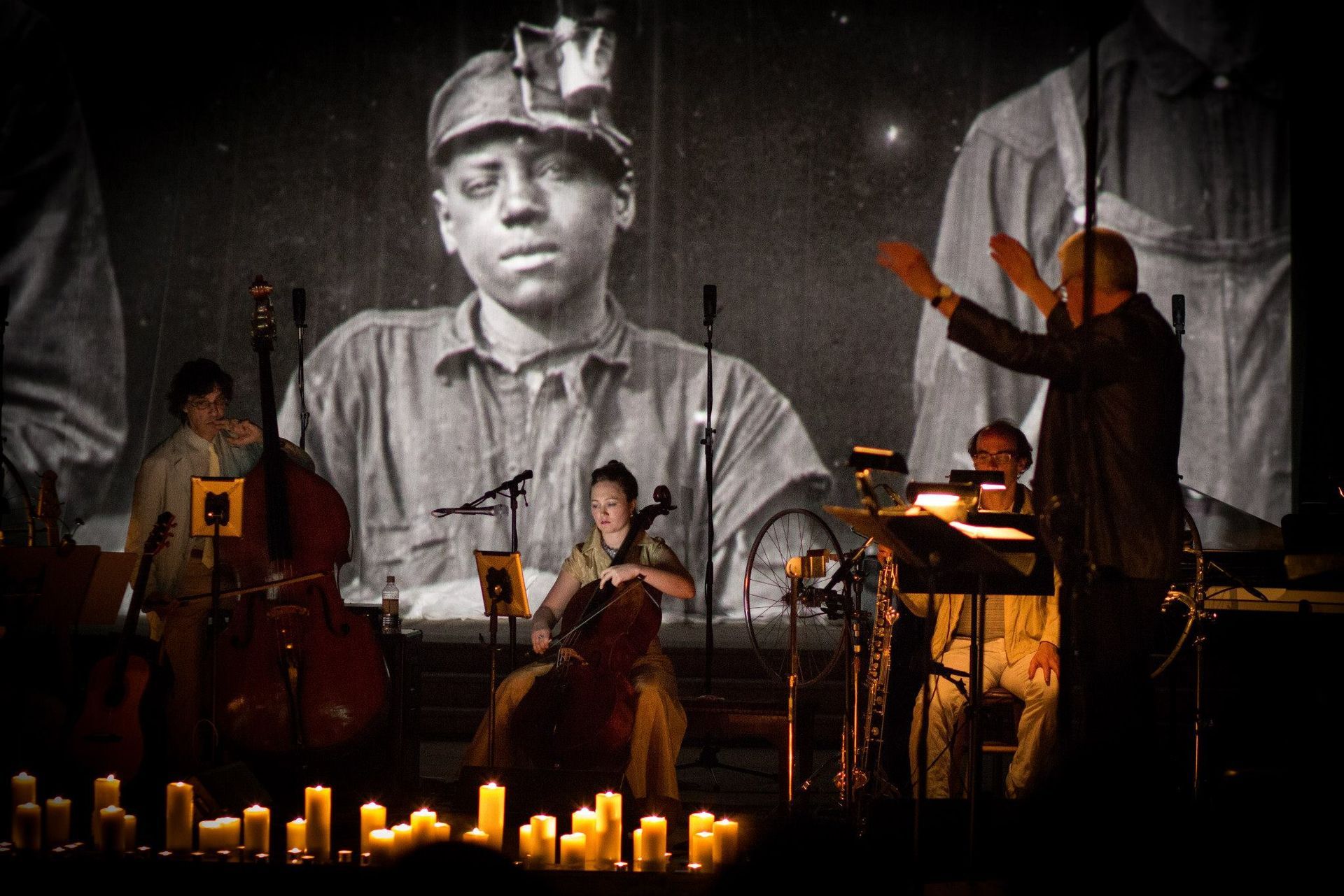 A performance with musicians and conductor. A black and white photo of a young miner is projected behind them, with candles in front.