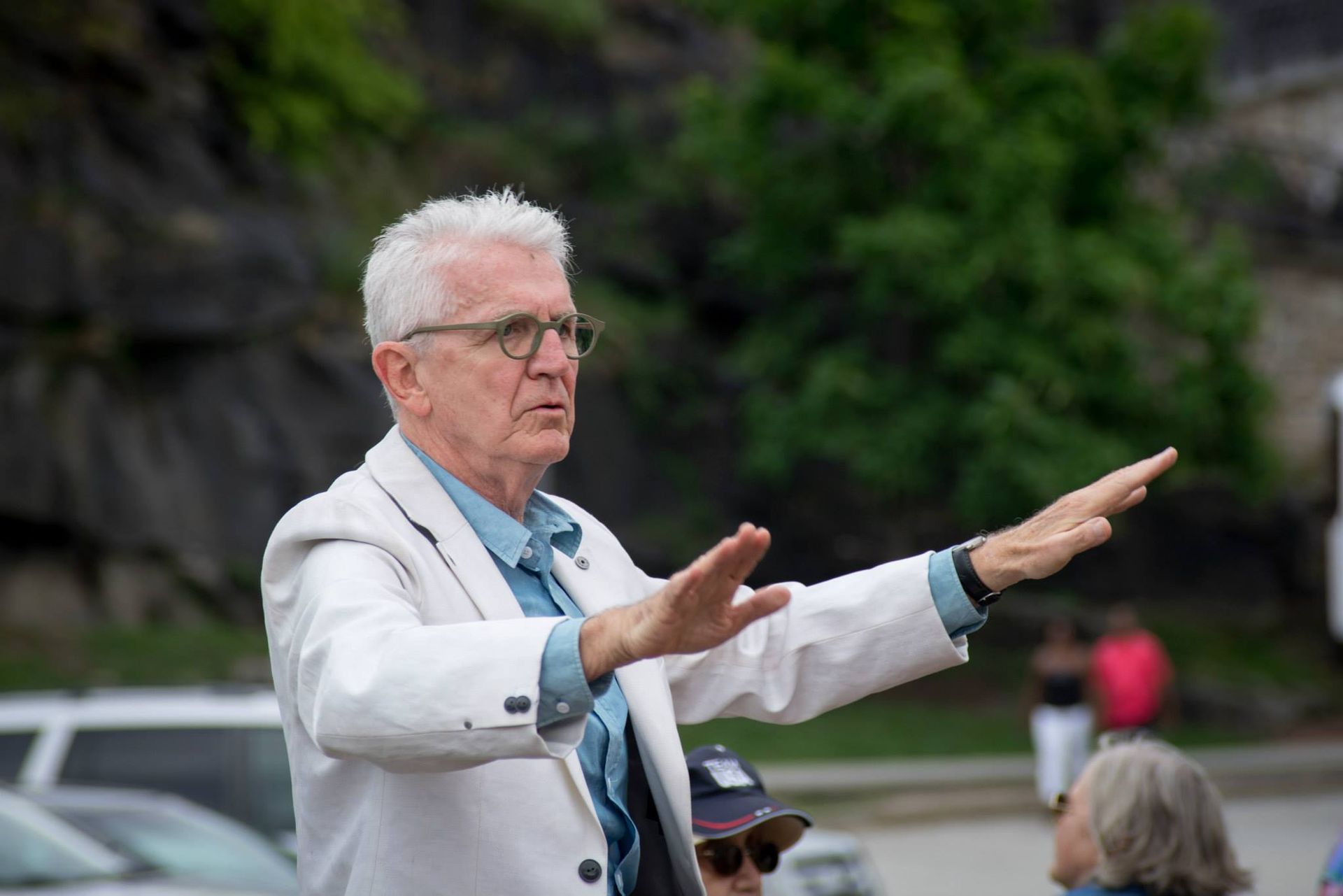 Man in white jacket gestures with hands open. Outdoors near a rocky backdrop.
