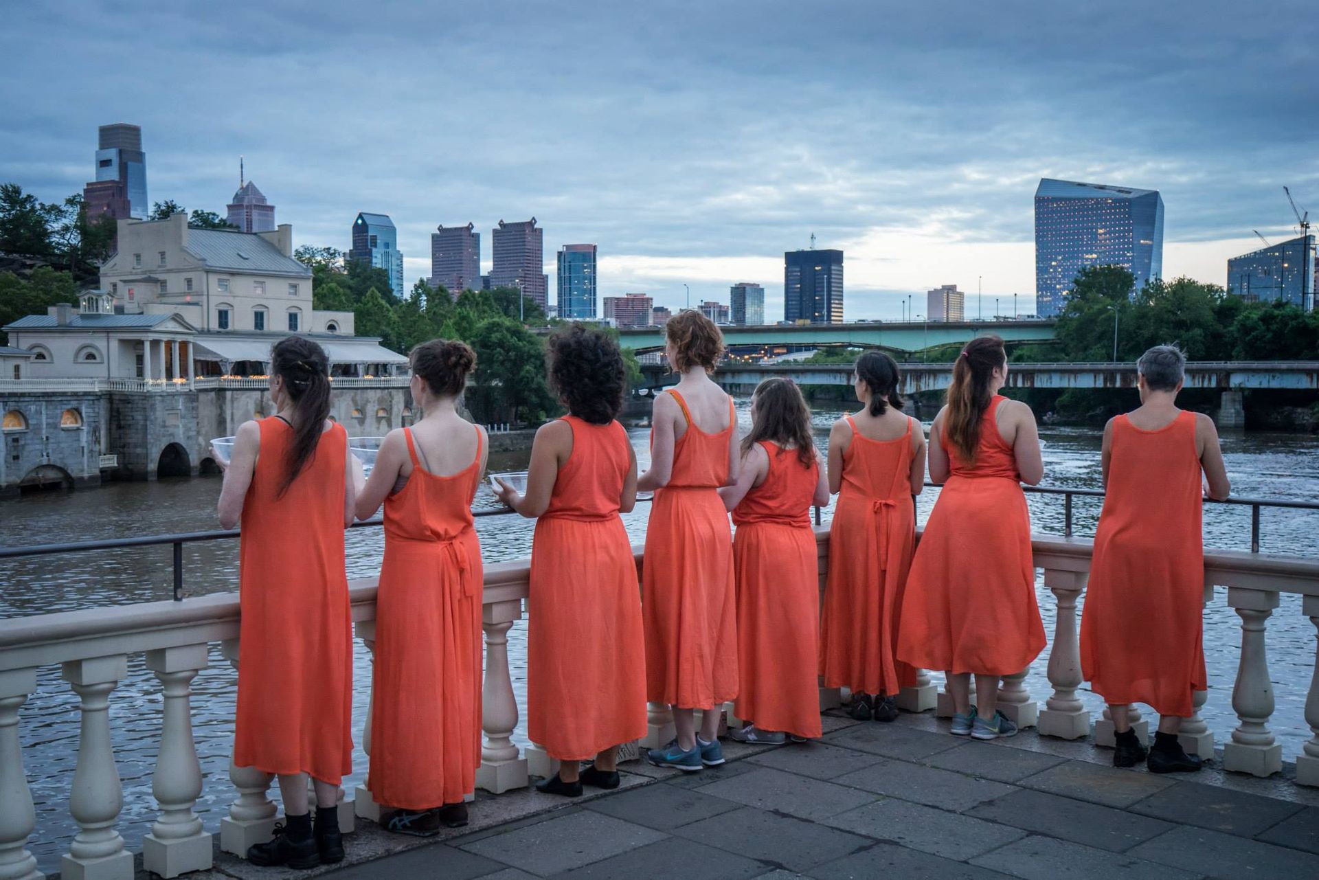 A group of women in orange dresses are standing on a bridge overlooking a body of water.