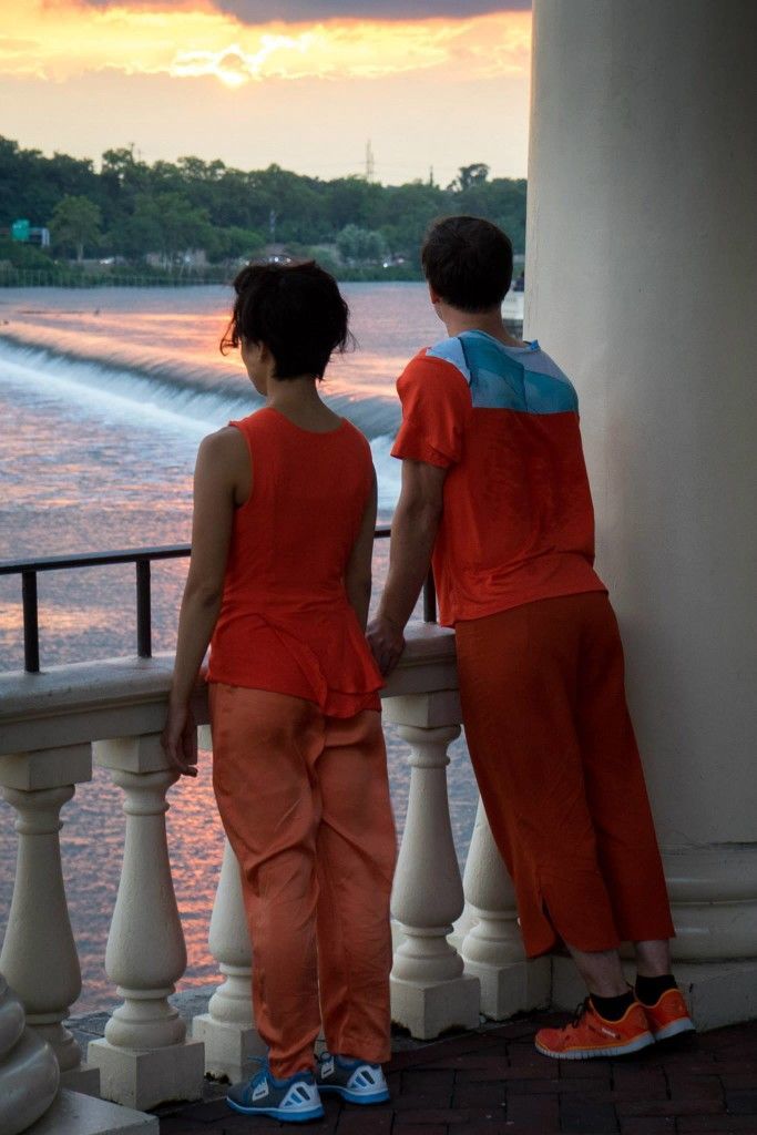 Two people are standing on a balcony holding hands and looking at the water.