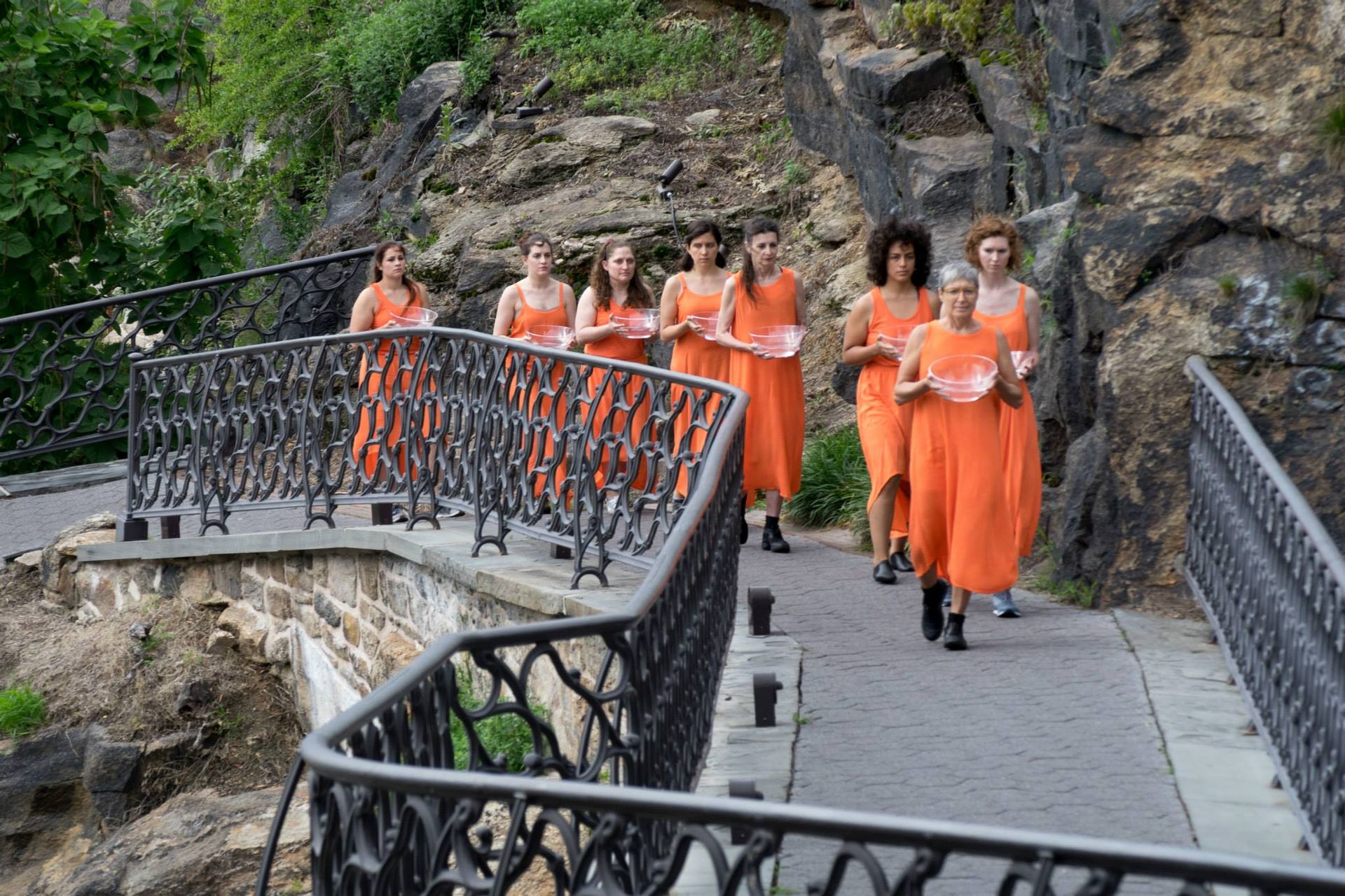 A group of women in orange dresses are walking across a bridge.