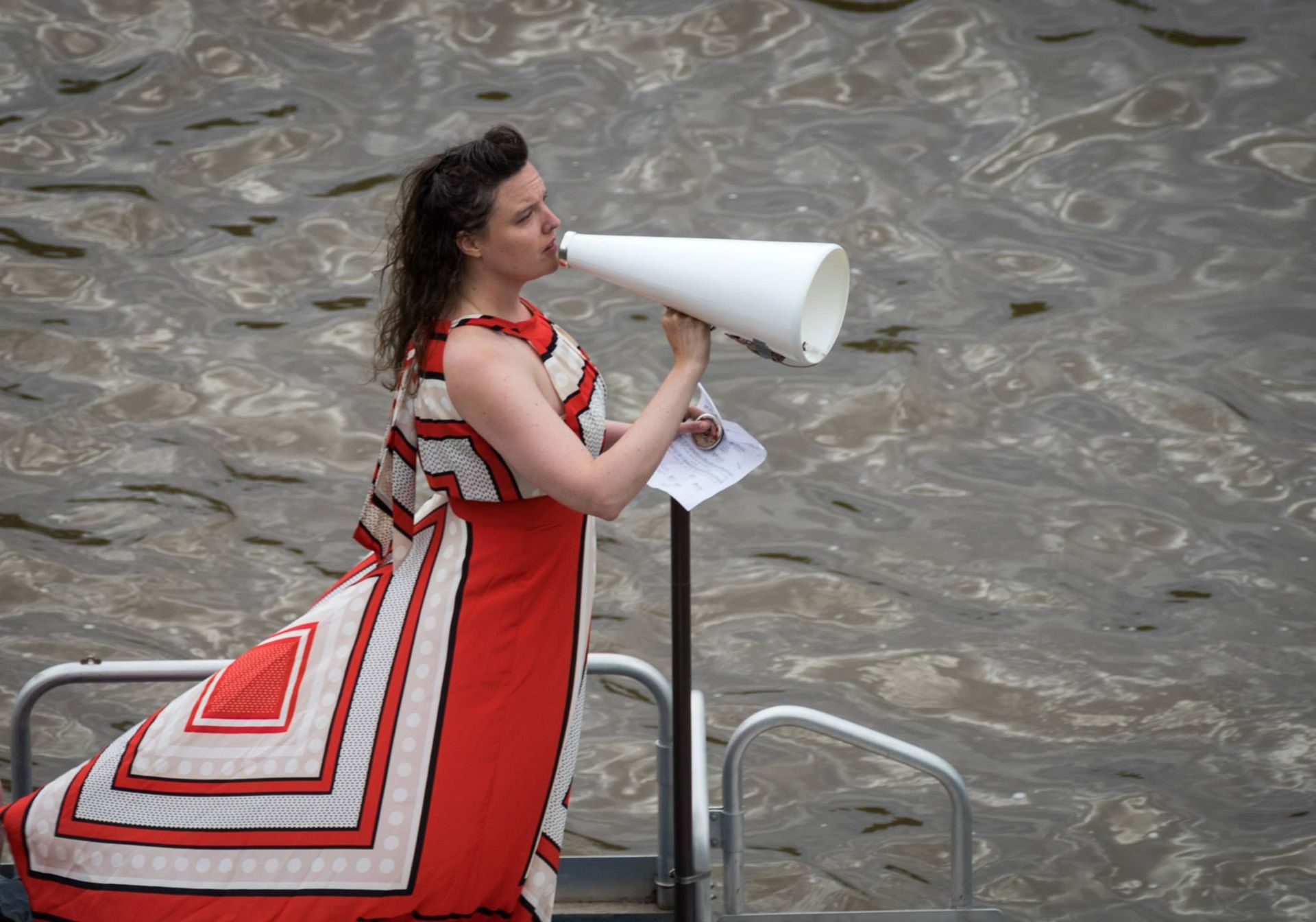 A woman in a red and white dress is holding a megaphone in front of a body of water.