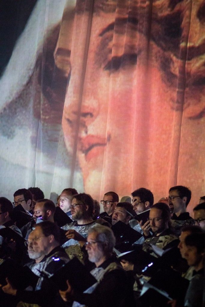 A large group of people are sitting in front of a large painting of a woman 's face.