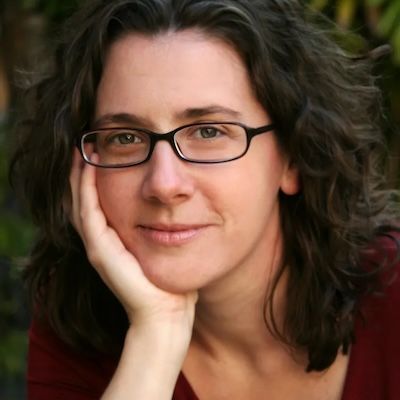 Person with glasses resting their chin on their hand, smiling softly outdoors.