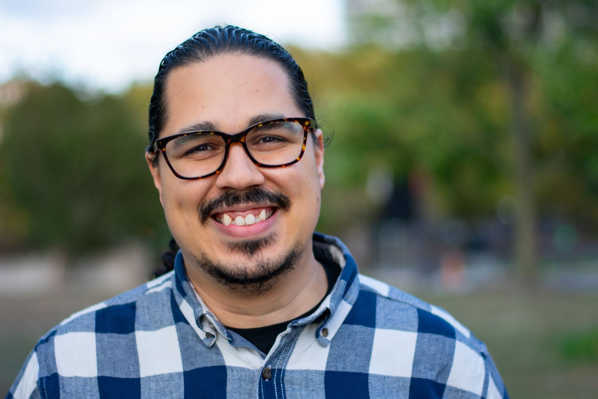 A man with long wavy dark hair in a blue tshirt and glasses standing in front of the Golden Gate Bridge wearing glasses smiling at the camera
