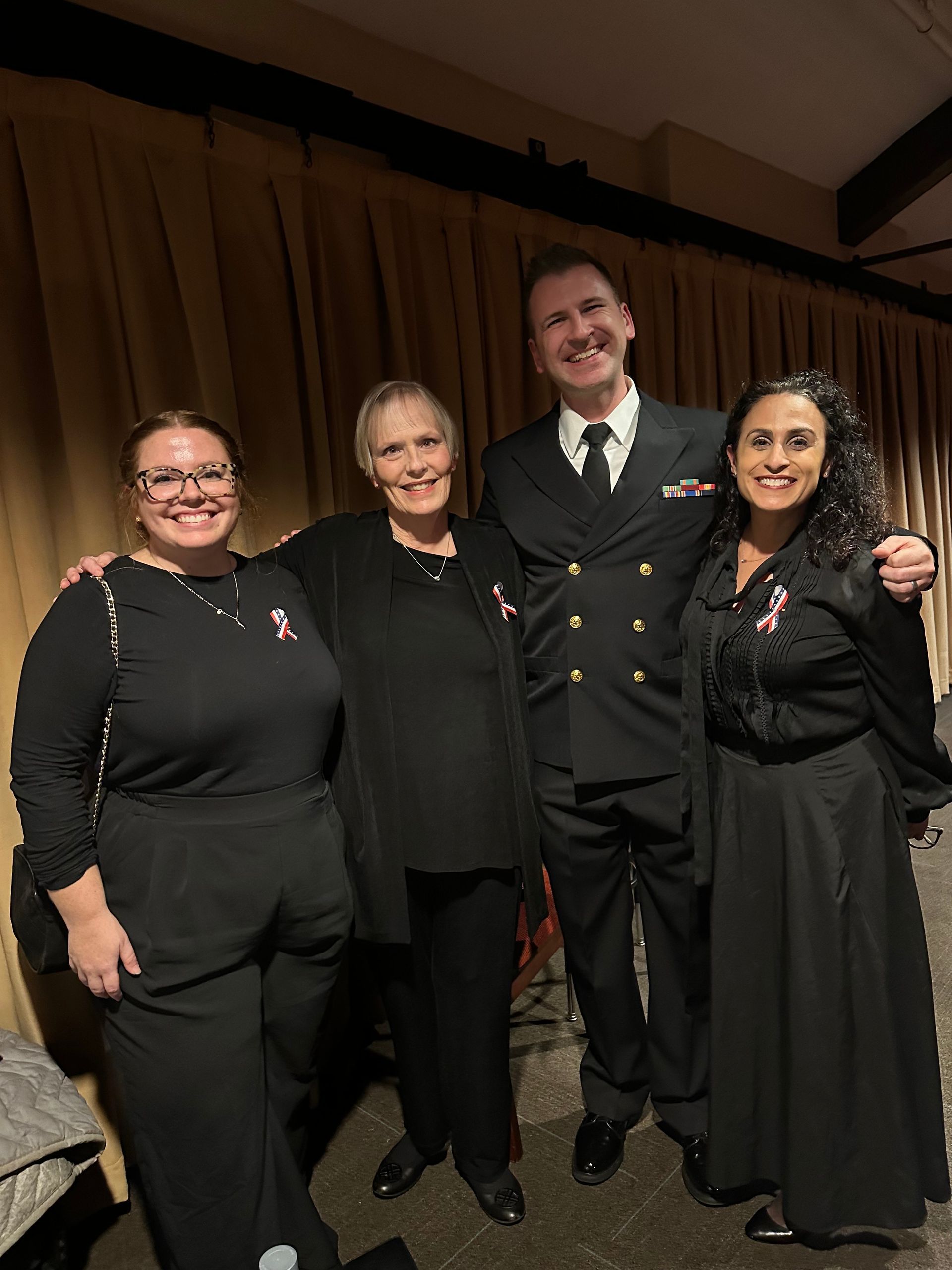 Four people in formal attire. Two women wear pink ribbons. One man in navy uniform. They pose indoors.