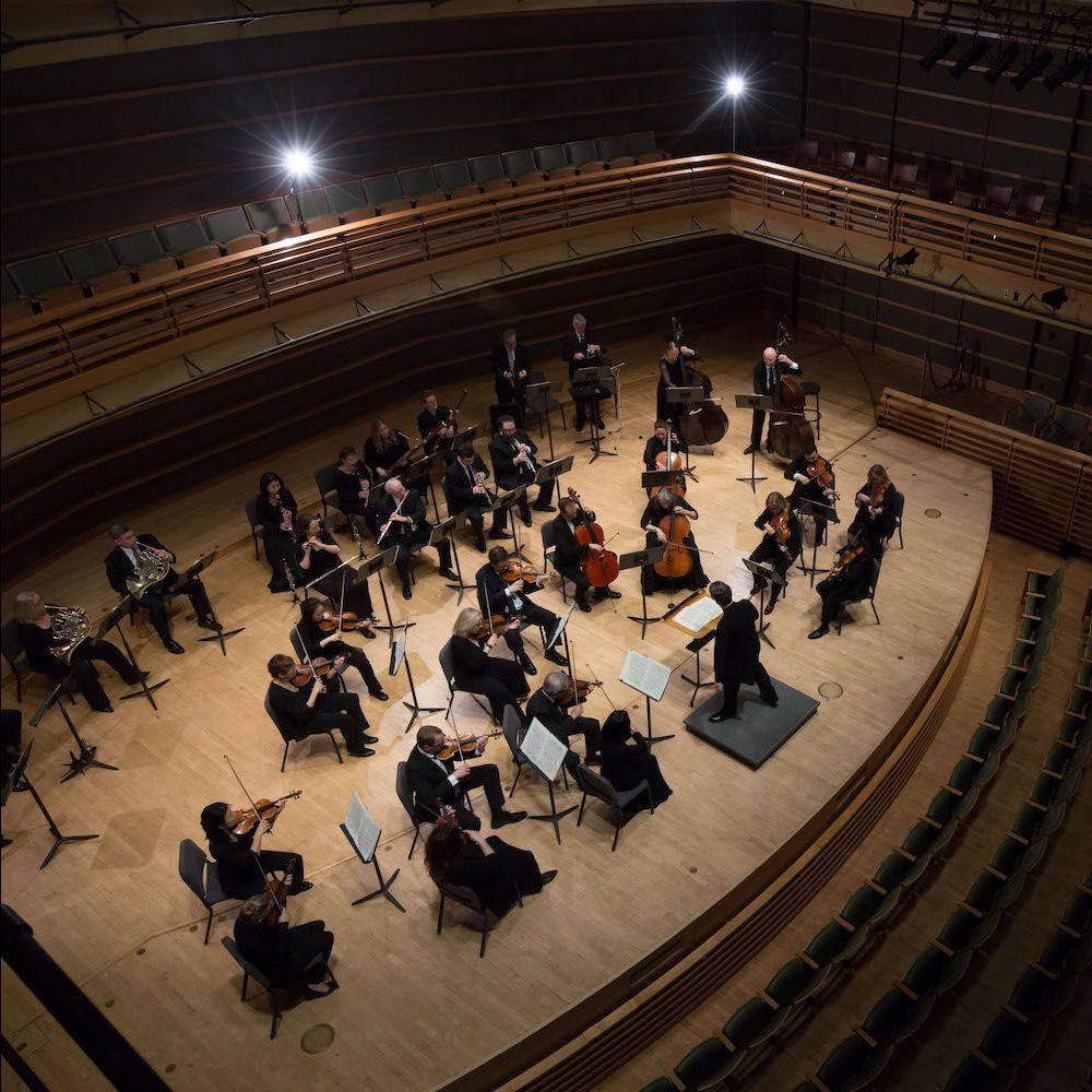Orchestra rehearsing on a wooden stage in a concert hall, viewed from above.