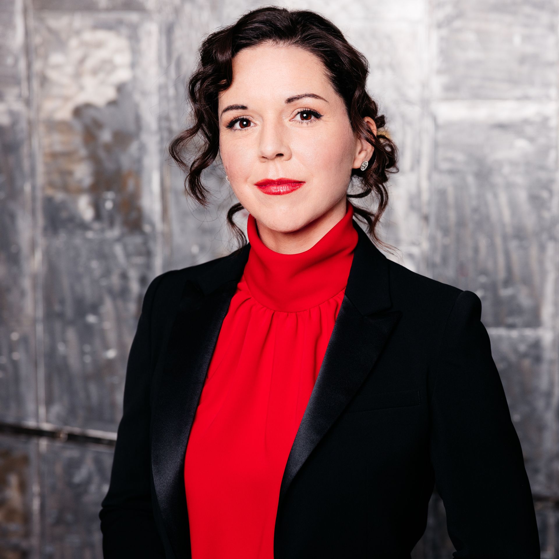 A woman in a black jacket and a red blouse standing in front a gray brick wall.