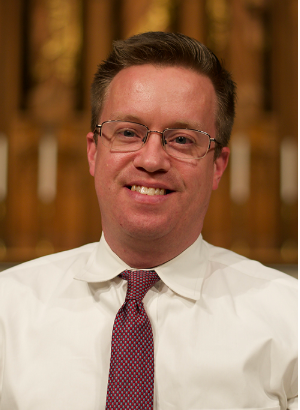 Portrait of a smiling man in glasses, white shirt, and maroon tie against a blurred indoor background