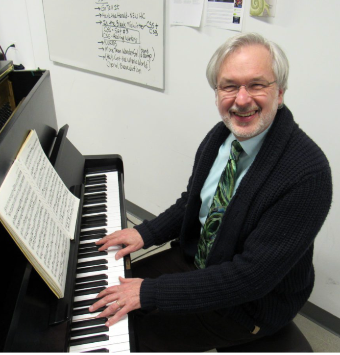 Smiling man playing piano in a classroom, with sheet music on the stand.