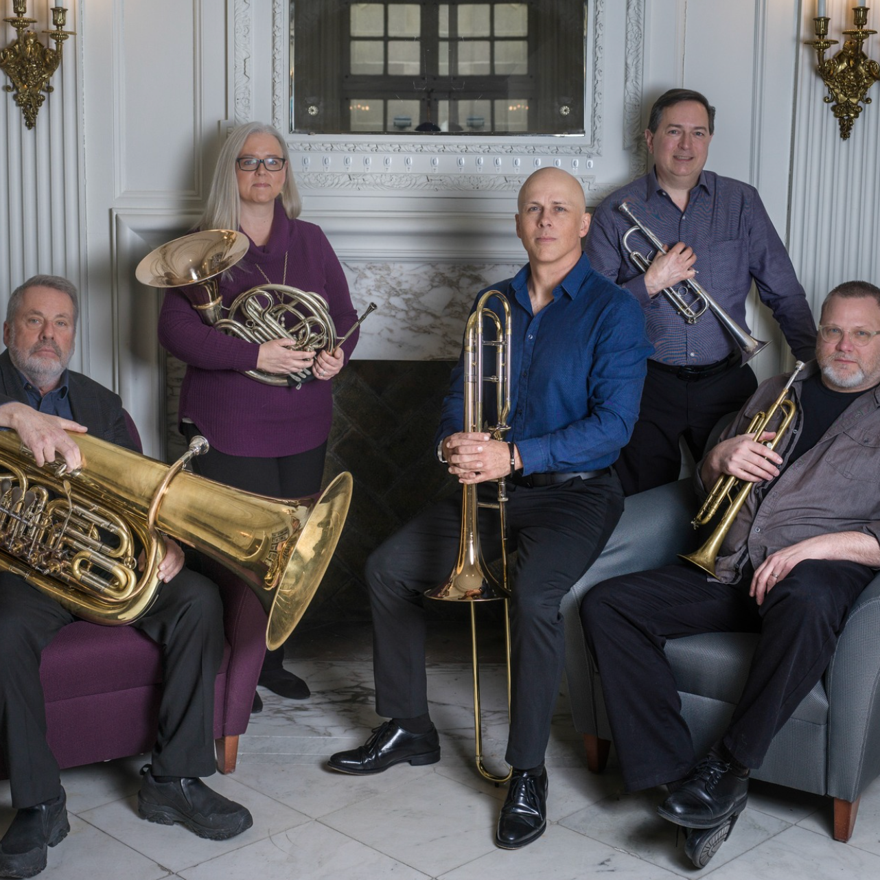 Five musicians posing indoors with brass instruments, including tuba, French horn, trombone, and trumpet.