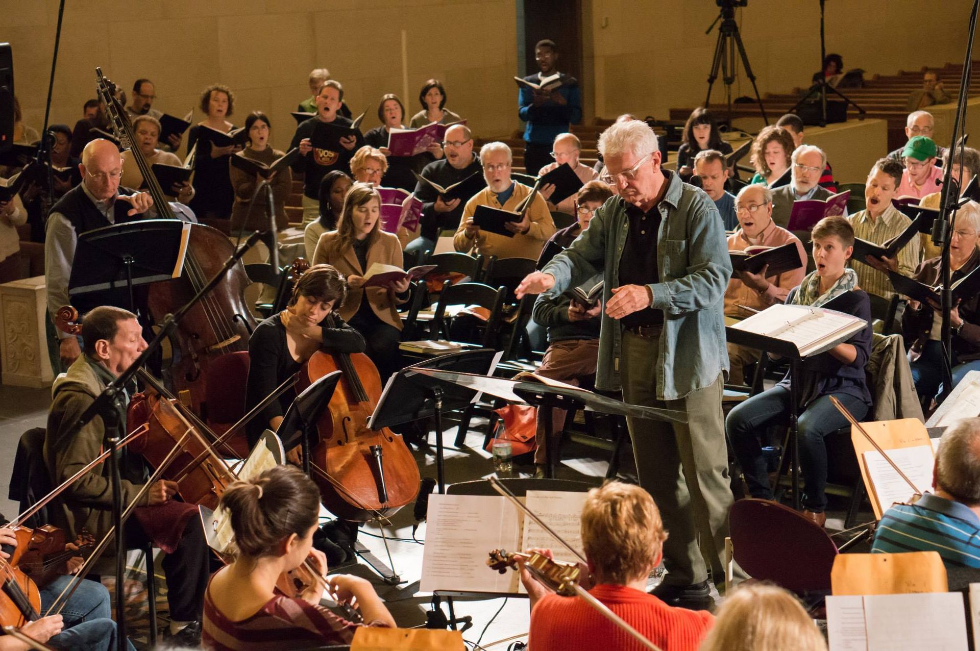 Orchestra rehearsing; conductor leads, musicians with instruments and sheet music, choir in background, concert hall.