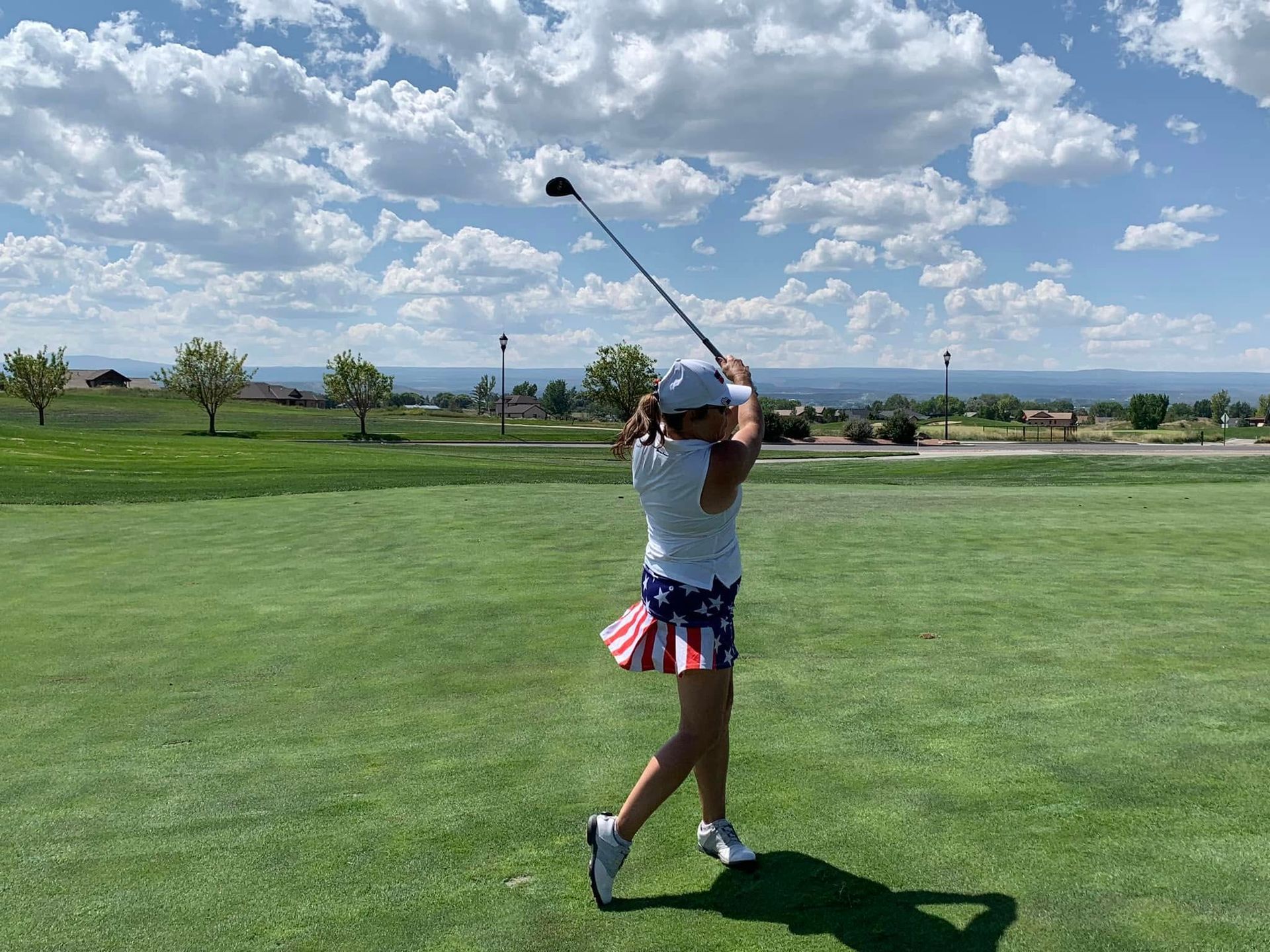 Woman in patriotic outfit swinging a golf club on a green course, blue sky with clouds in background.