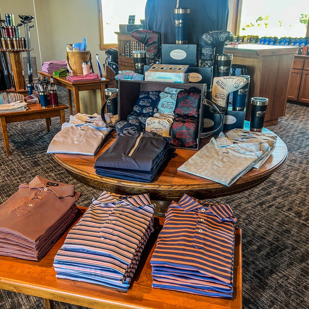 Golf shop display of shirts, drinkware, and accessories on round and rectangular tables.