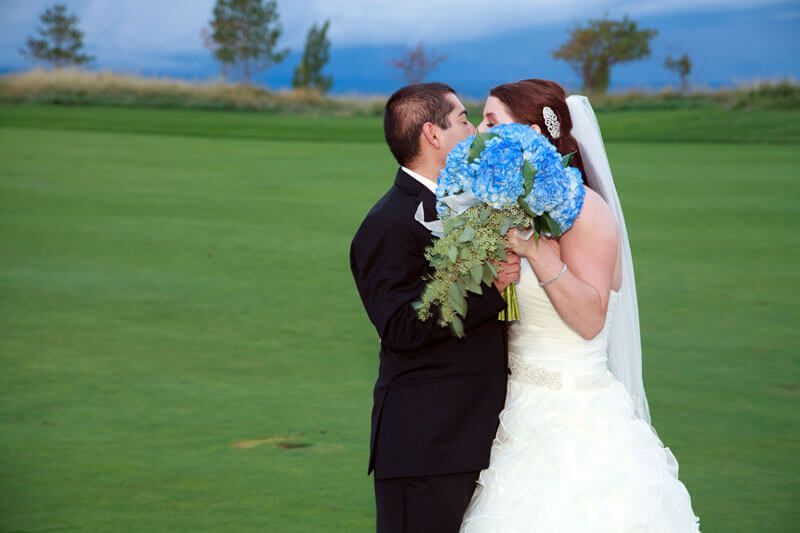Bride and groom kissing, partially obscured by blue hydrangea bouquet, on green golf course.