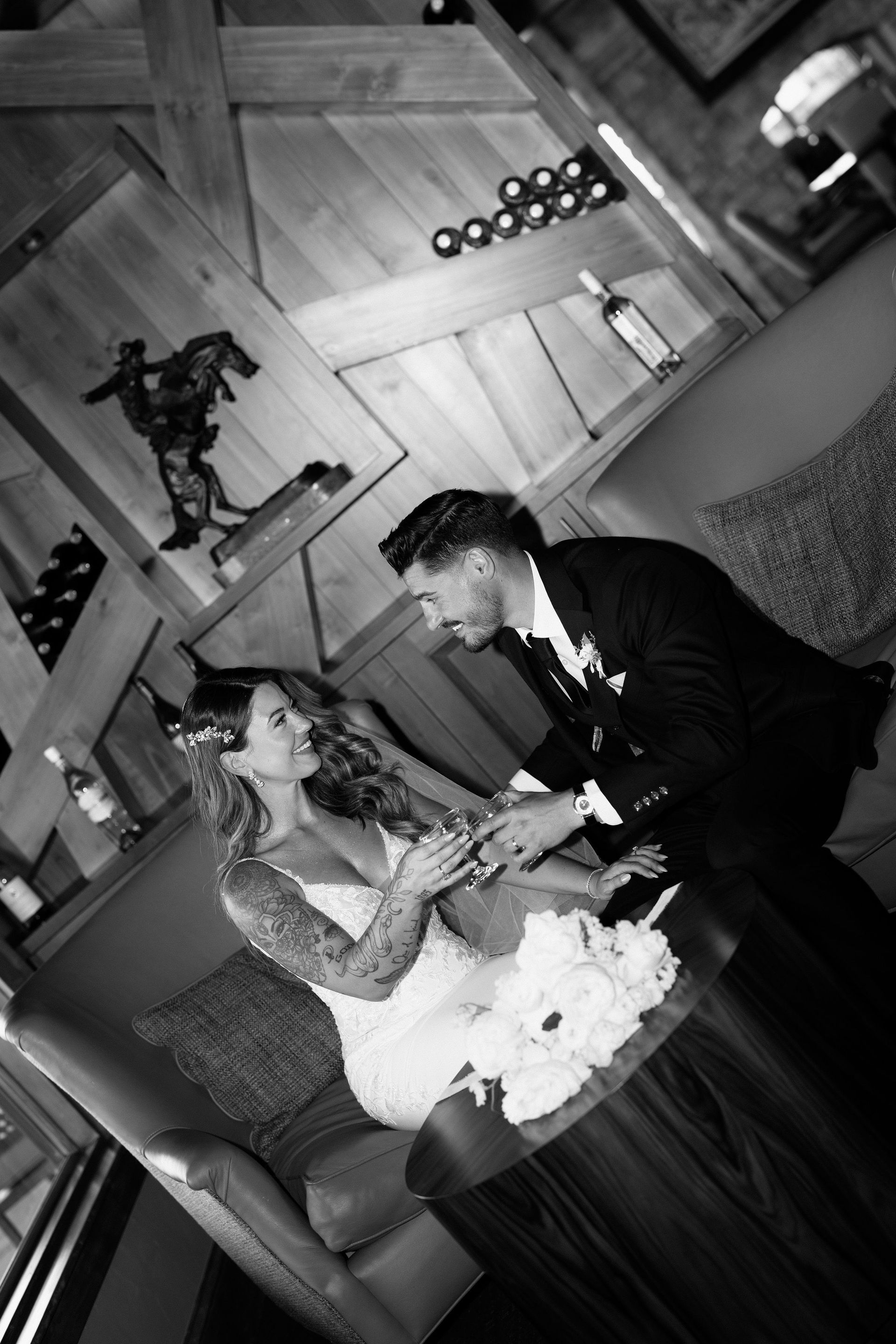 Bride and groom sit, hands clasped, in a room with wood paneling, bottles of wine on display.