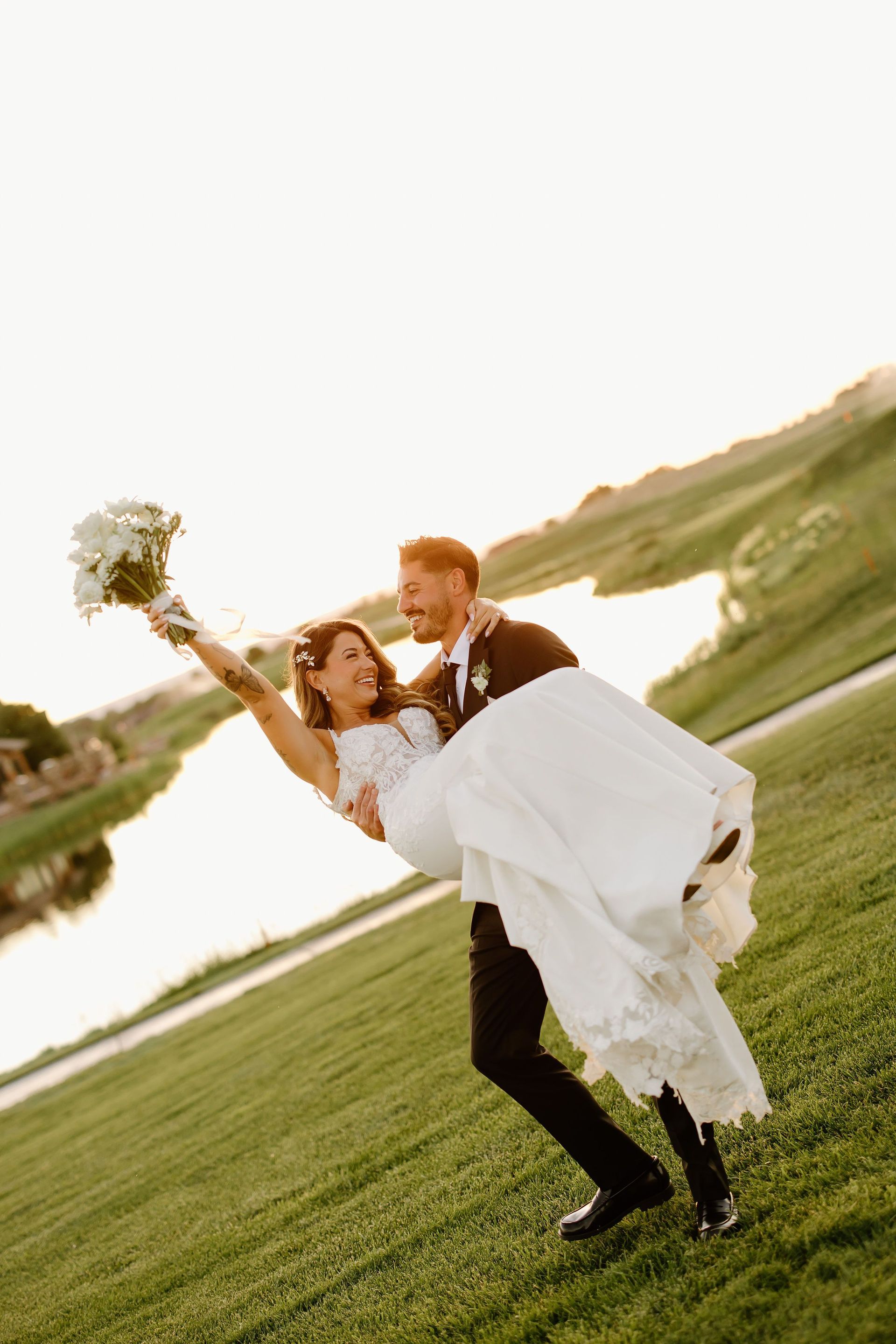 Groom carrying bride, both smiling and celebrating. They are on a green lawn near water at sunset.