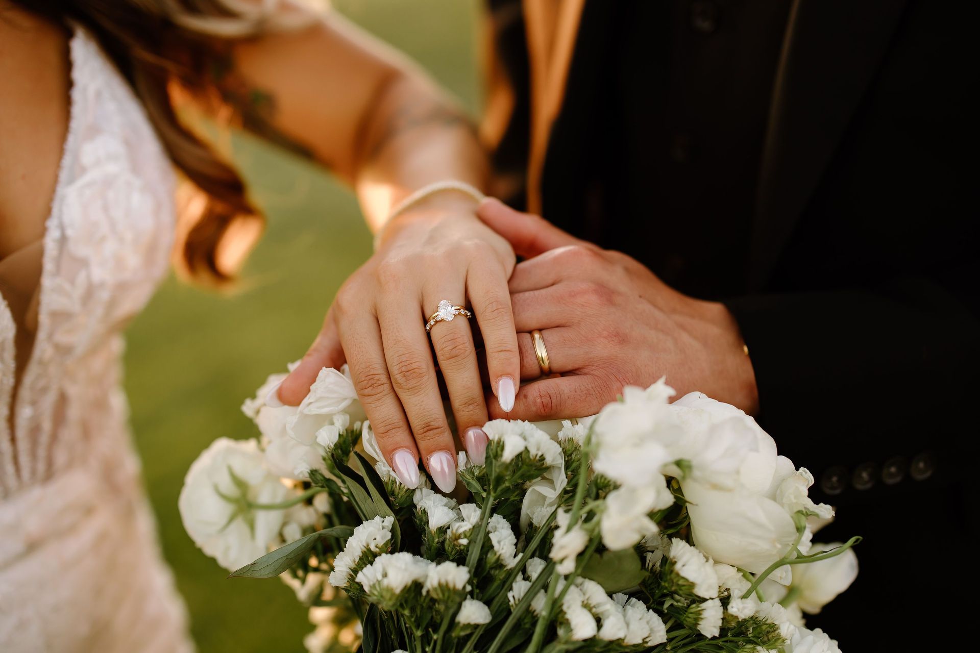 Couple's hands clasped, showing rings, over a white floral bouquet. Groom's black suit contrasted with bride's dress.
