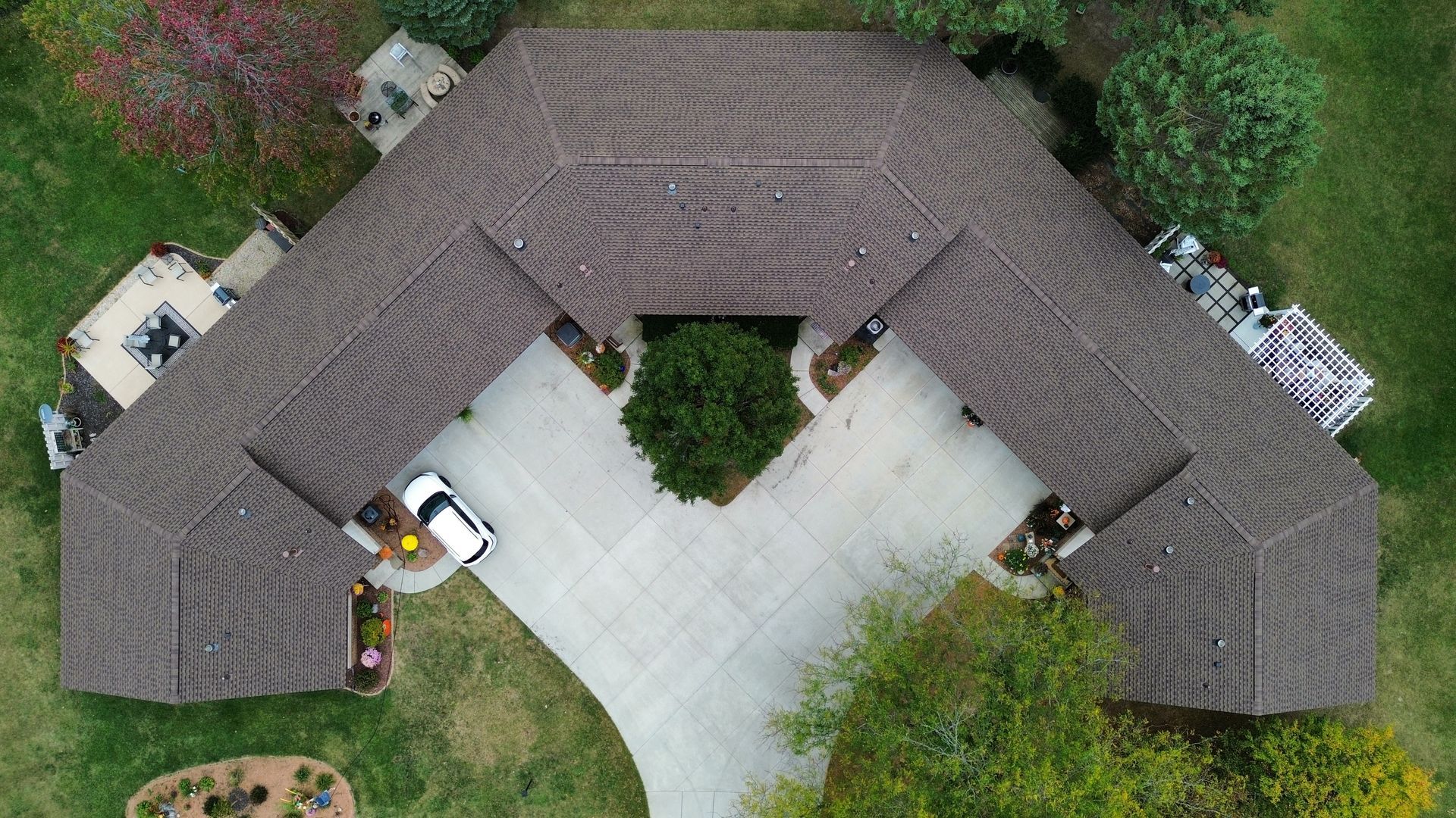 A high-angle aerial view of a U-shaped house with a dark brown shingled roof, a paved driveway, and surrounding lawn.