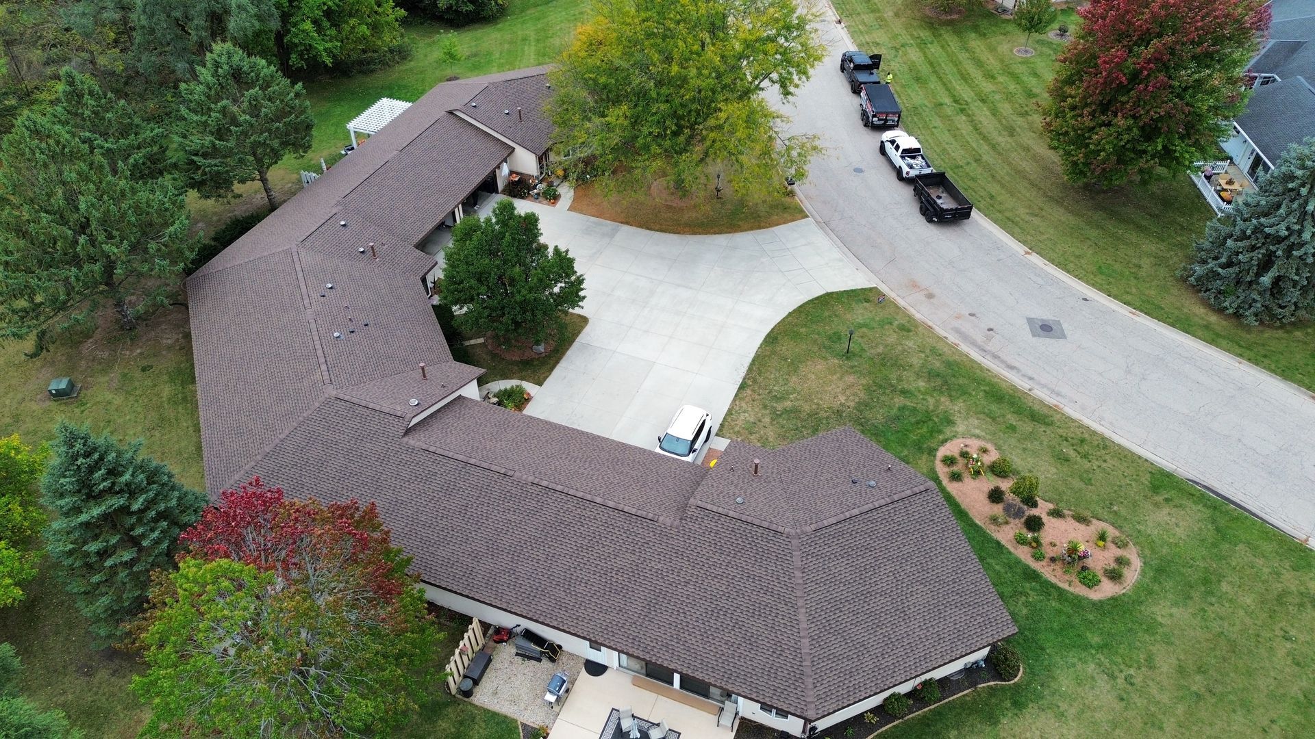 Aerial view of an L-shaped house with a dark brown shingled roof, a paved driveway, and surrounding green lawn and trees.