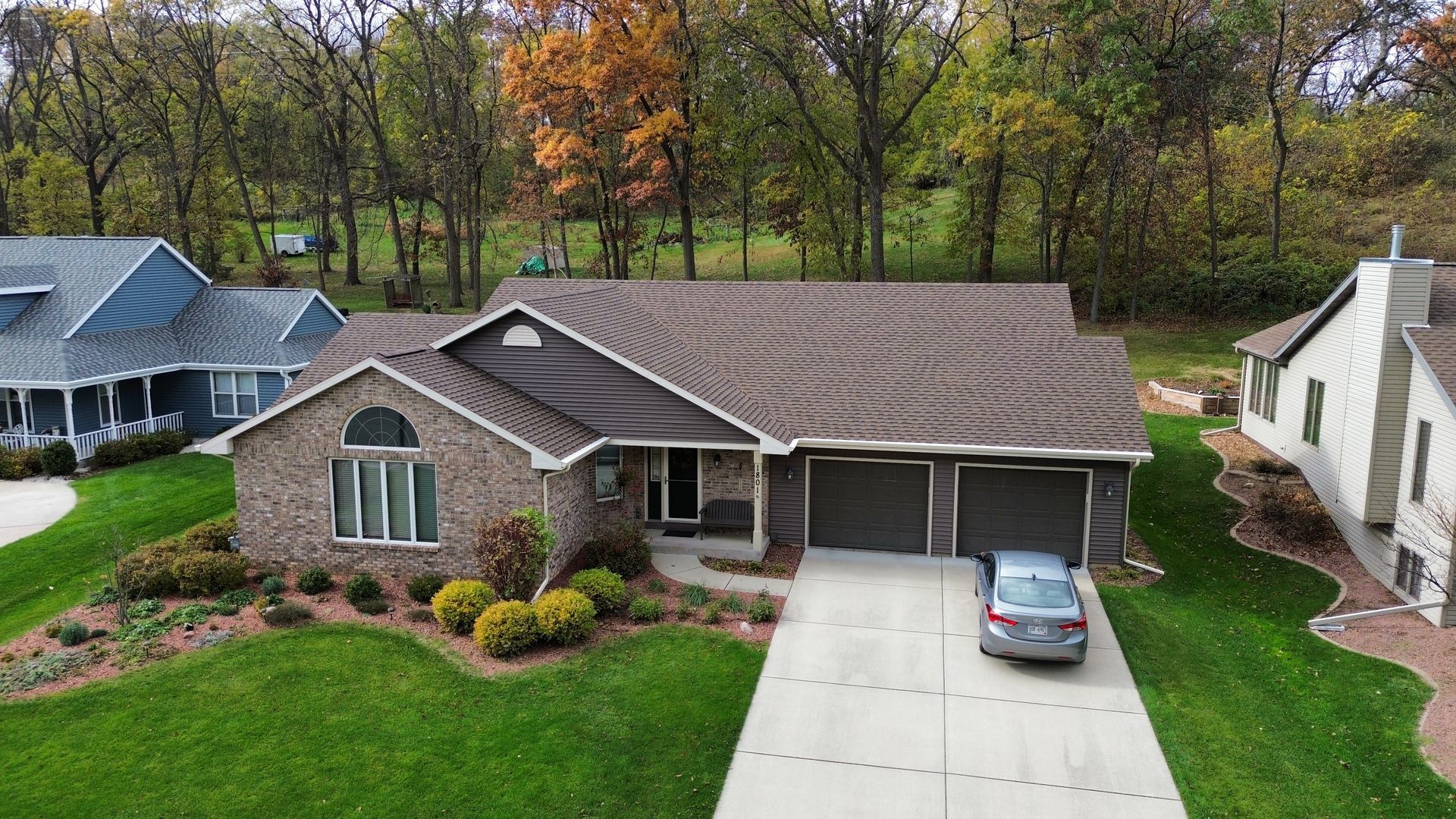 An aerial view of a brick house with a brown roof, a concrete driveway with a silver car, and a green lawn.