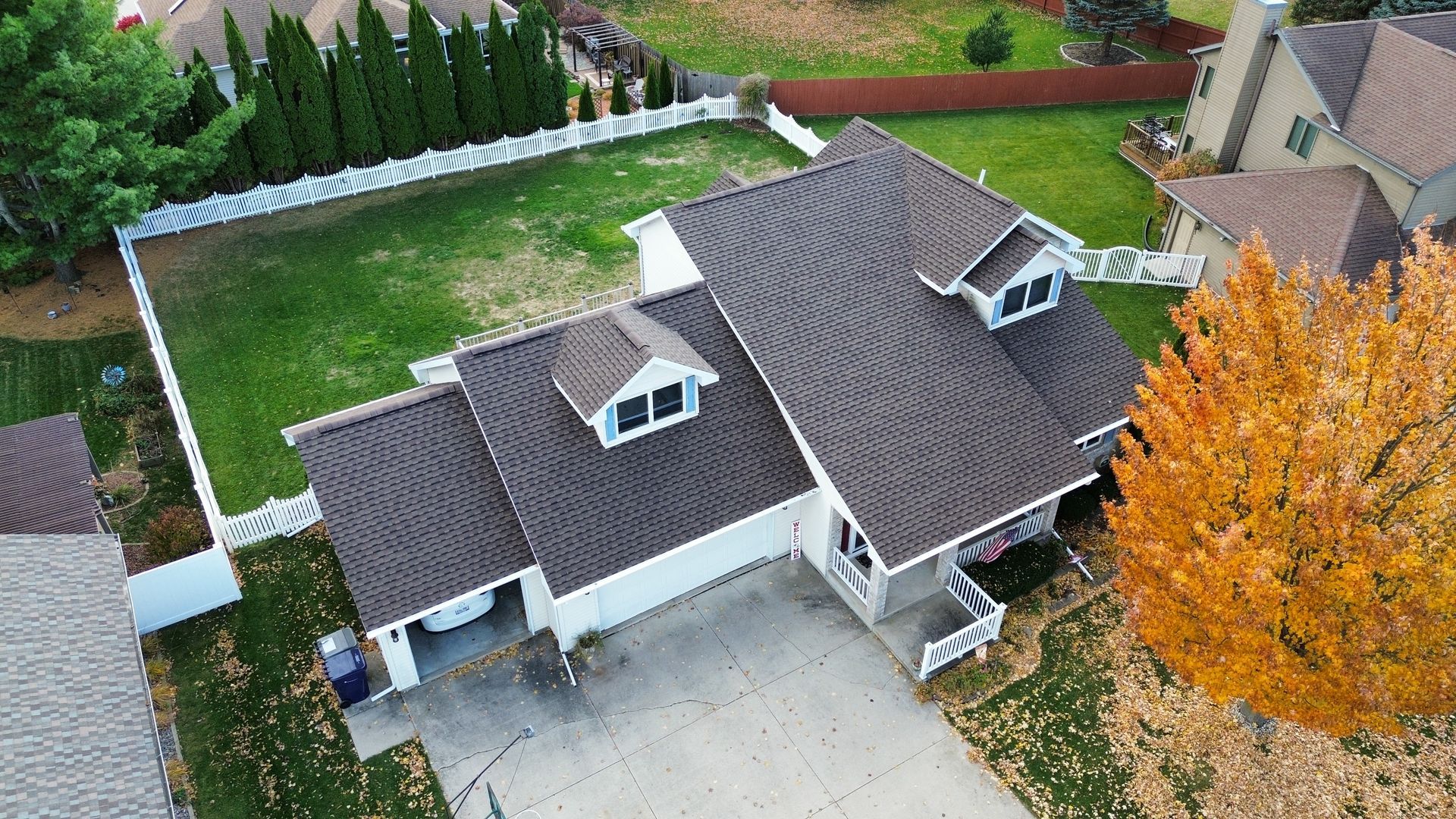 An aerial view of a house with a dark brown shingled roof, a paved driveway, a green lawn, and a vibrant orange tree.