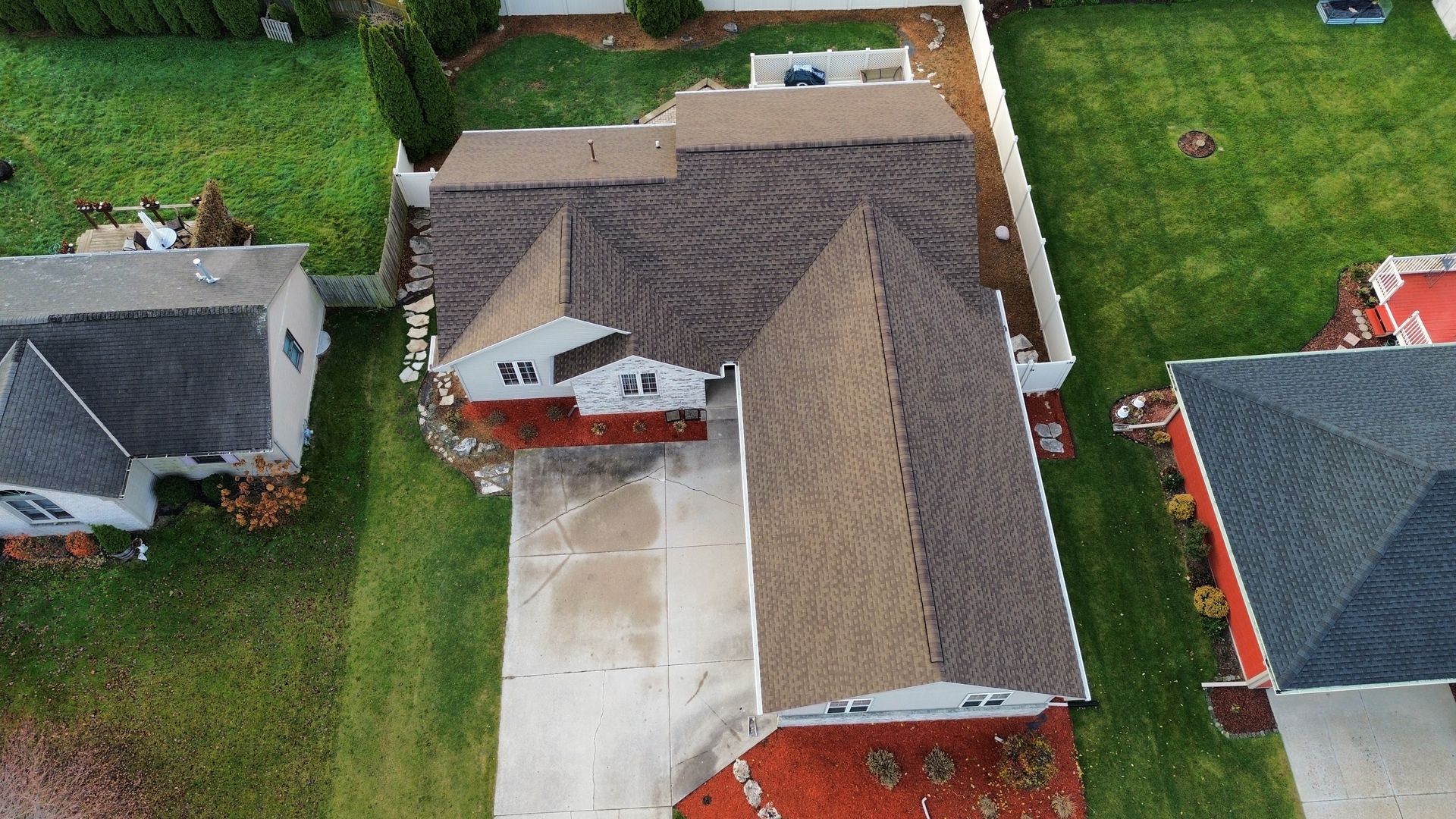 Aerial view of a residential house with a brown shingled roof, a paved driveway, and surrounding green lawn.