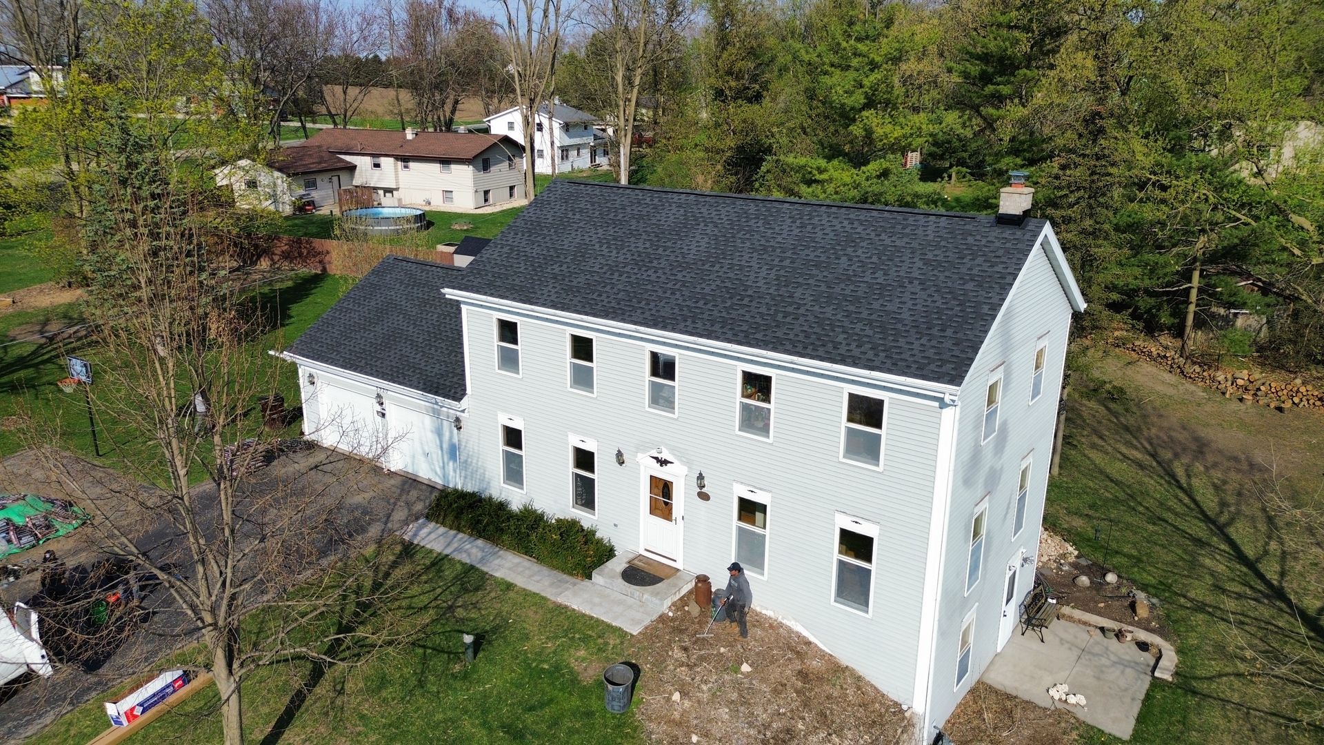 An aerial view of a two-story light gray house with a new dark gray shingled roof, surrounded by a lawn and trees.