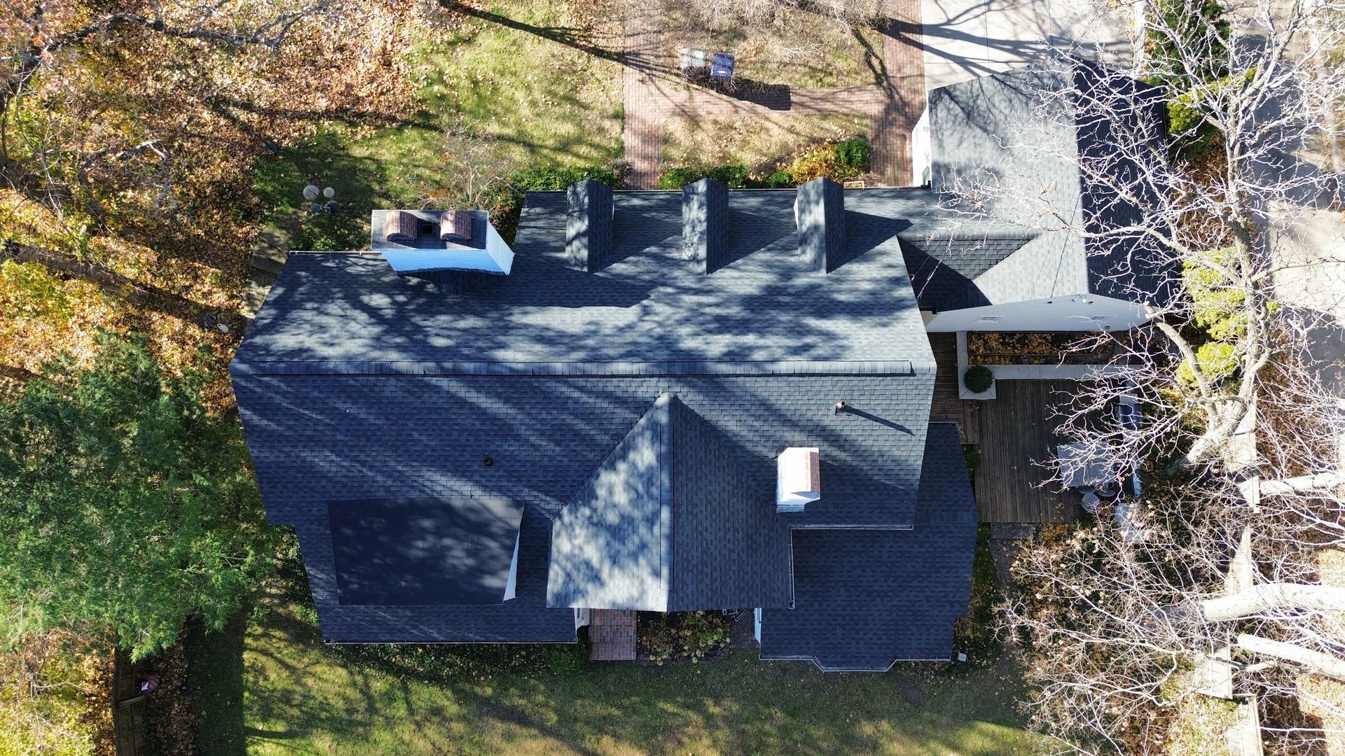 An aerial view shows a dark-roofed residential house surrounded by trees on a sunny day.