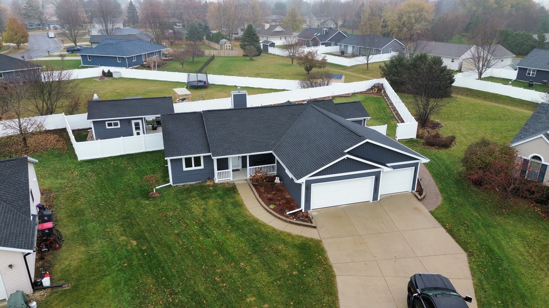 An aerial view of a dark gray house with a white garage and white fence in a suburban residential neighborhood.