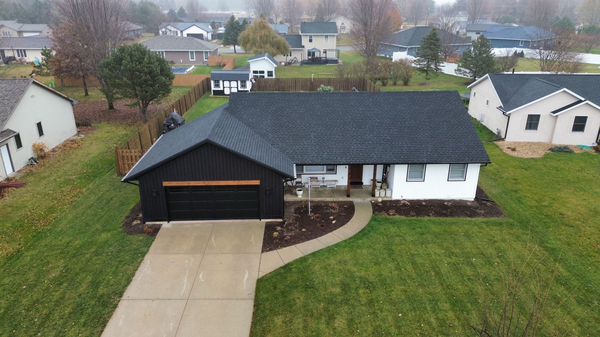 Aerial view of a white, one-story house with a black garage and roof, surrounded by a lawn in a neighborhood.