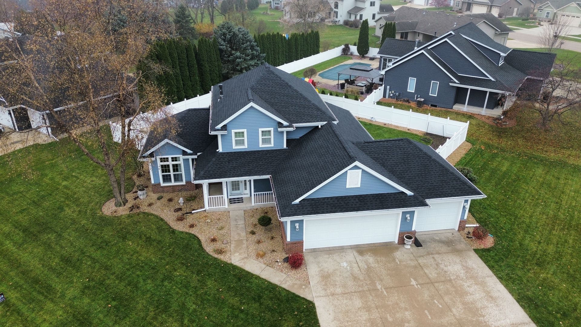 An aerial view of a blue two-story house with a white garage, dark roof, and a fenced backyard with a pool.