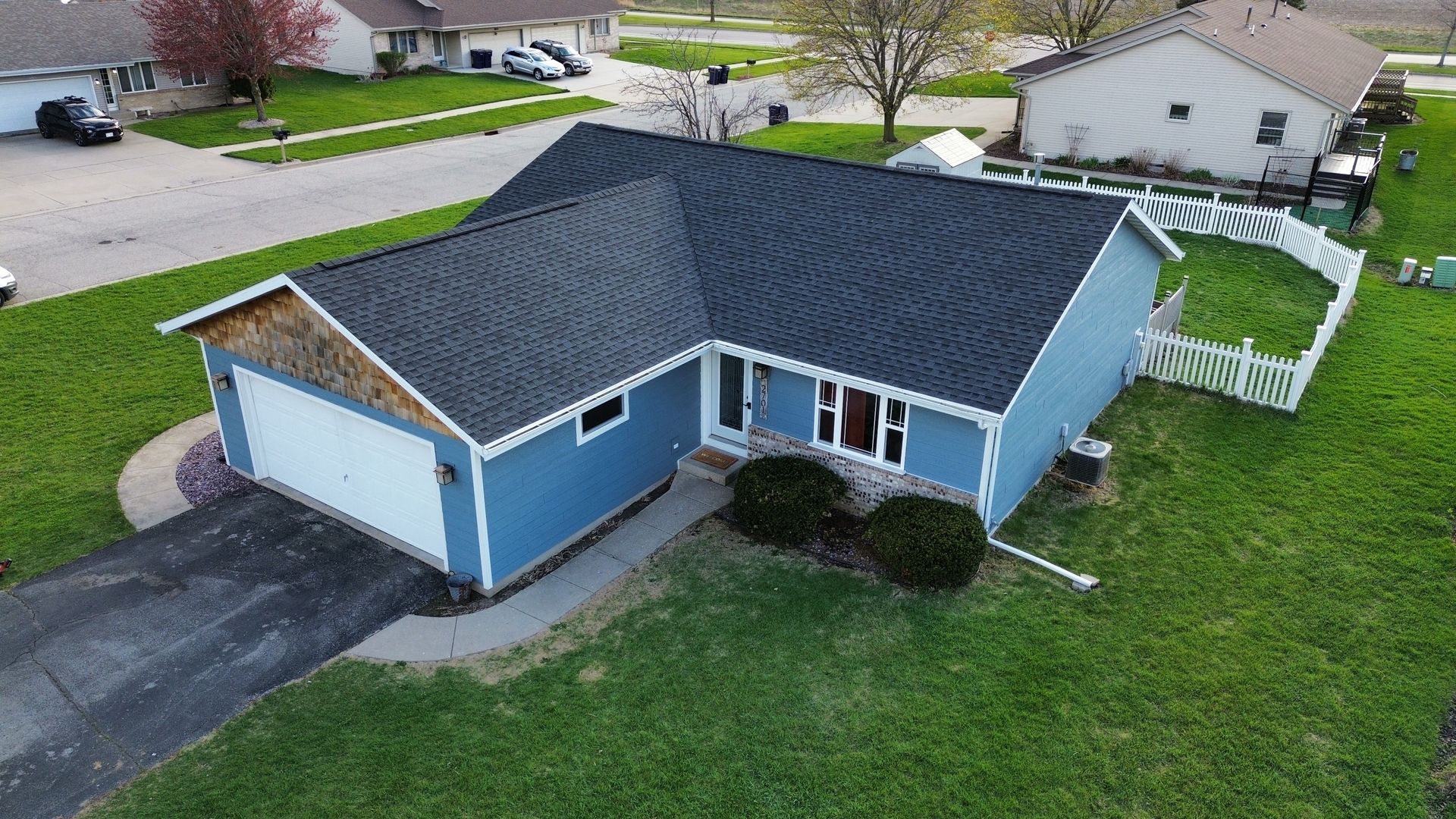 An aerial view of a blue, single-story house with a black roof, a white garage door, and a white fence in the yard.