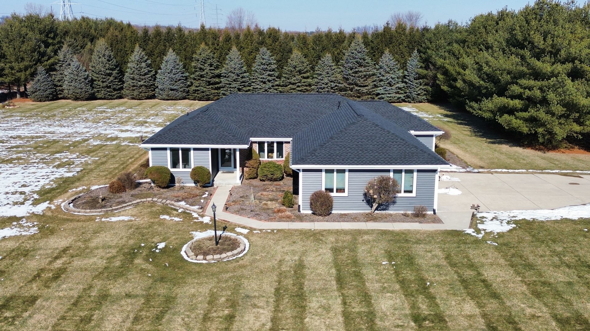 An aerial view of a gray, single-story house with a dark shingled roof, surrounded by lawn and a row of evergreen trees.
