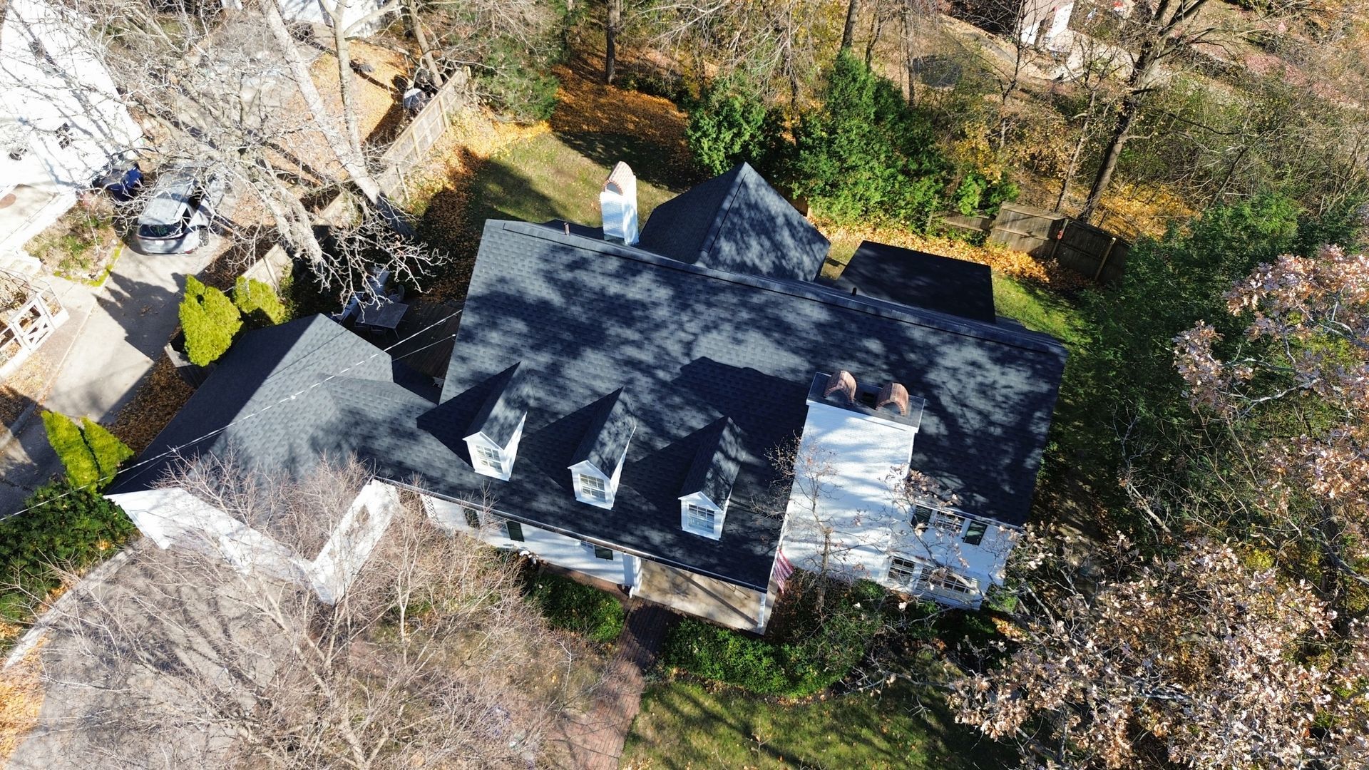 An aerial view of a dark-roofed suburban house with three dormer windows, surrounded by trees and a lawn.